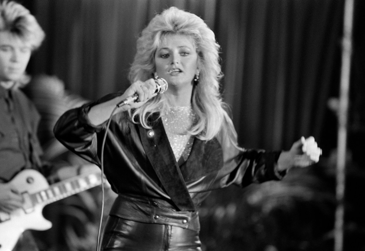 Black and white photo of a blonde woman with voluminous hair singing into a microphone in a leather outfit, with a guitarist visible behind her.