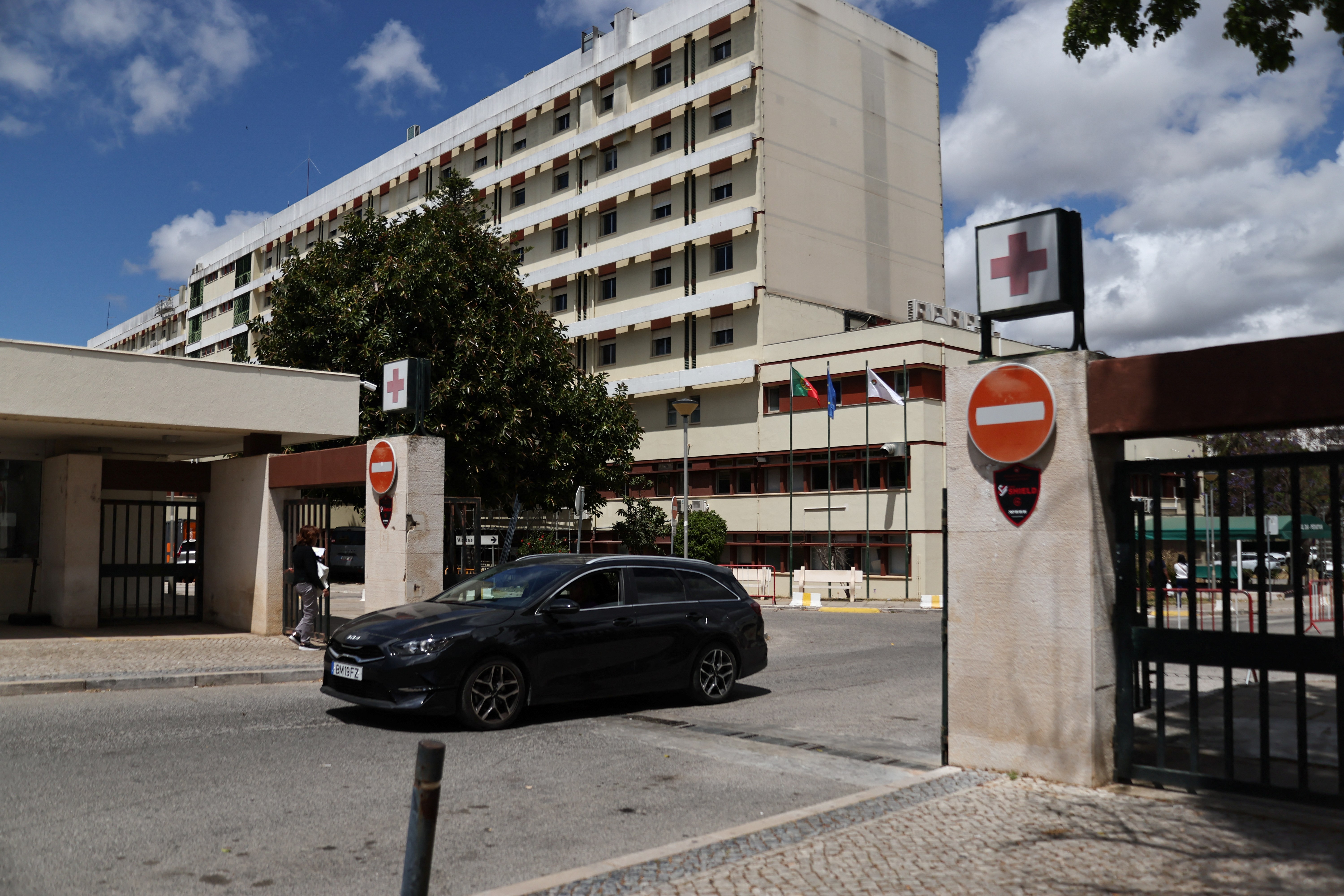 Click to open the image gallery Exterior of a multi-story hospital building with red cross signs at the entrance gate, a dark car passing through, under a partly cloudy sky.