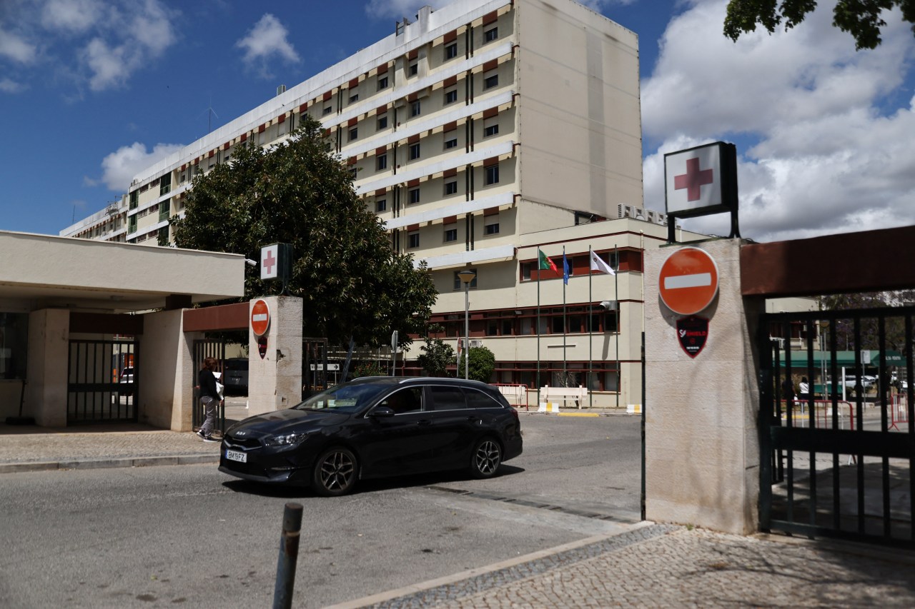 Exterior of a multi-story hospital building with red cross signs at the entrance gate, a dark car passing through, under a partly cloudy sky.