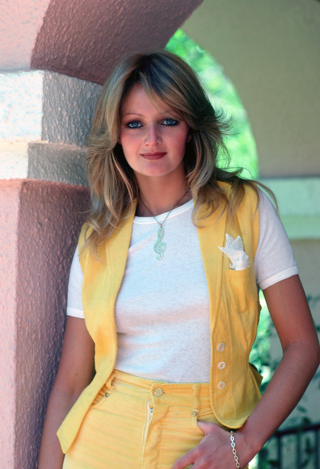 Young blonde woman with blue eyes wearing a yellow vest over a white t-shirt, posing near a stone archway outdoors.