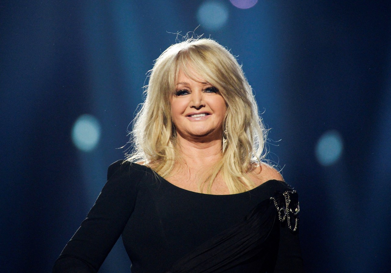 Blonde woman in a black off-shoulder dress smiling on a stage with blue bokeh lights in the background.