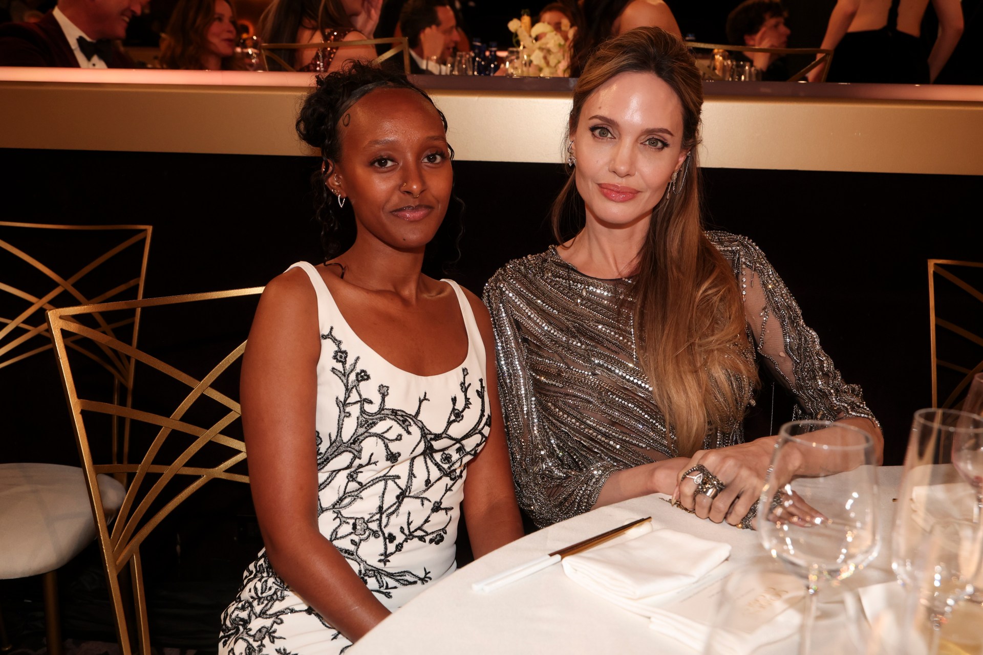 Zahara Jolie in a white floral dress and Angelina Jolie in a silver beaded gown sit together at a table at the Golden Globes.