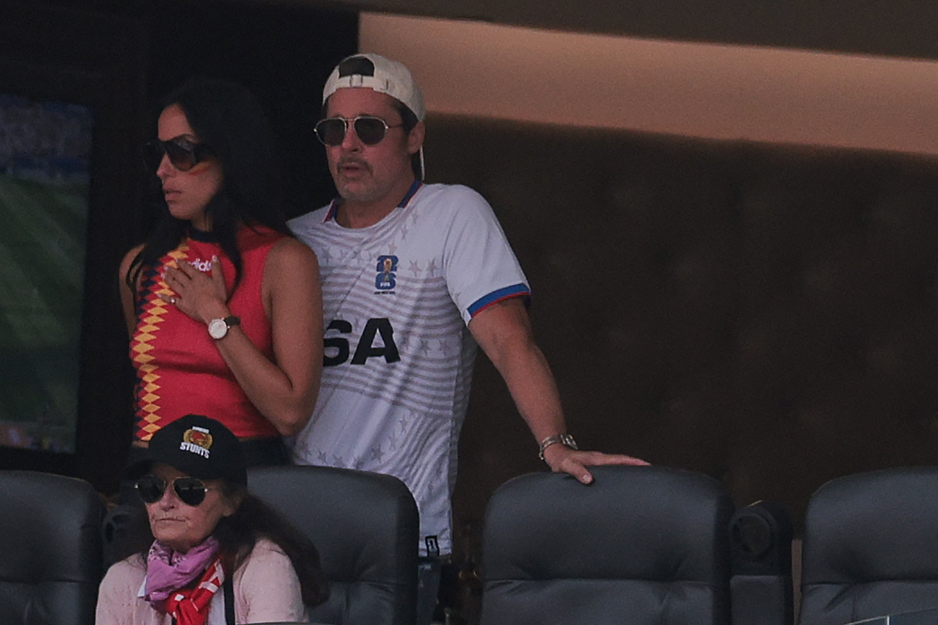 A man in a white USA jersey and backward cap and a woman in a red patterned top, both wearing sunglasses, stand together in stadium seating.