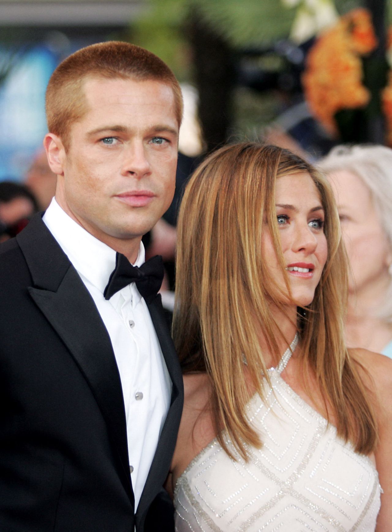 A man in a black tuxedo and bow tie stands beside a woman in a white dress with long highlighted hair on a red carpet.
