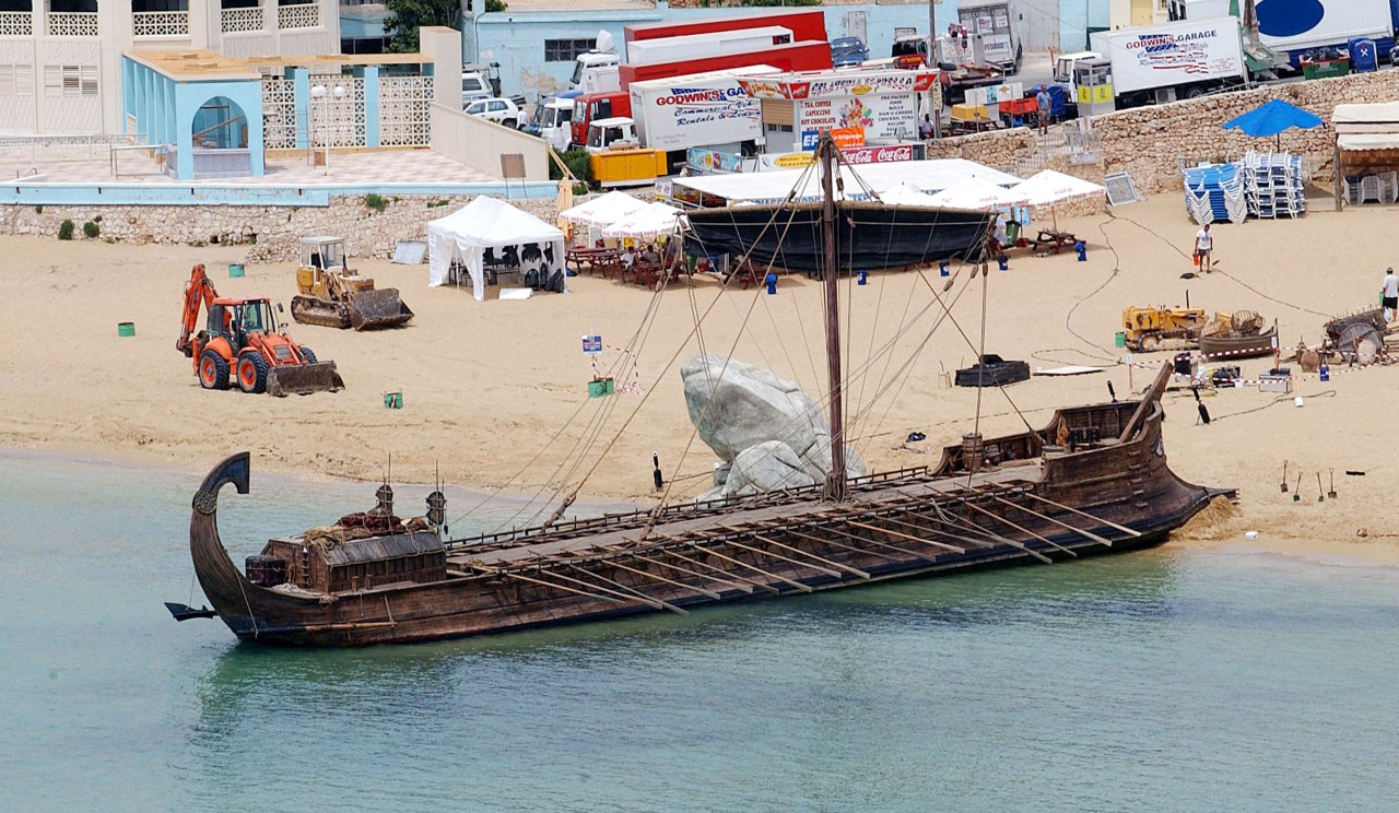 A large ancient-style wooden ship beached on sand near the water's edge, with construction machinery and tents on the beach behind it.