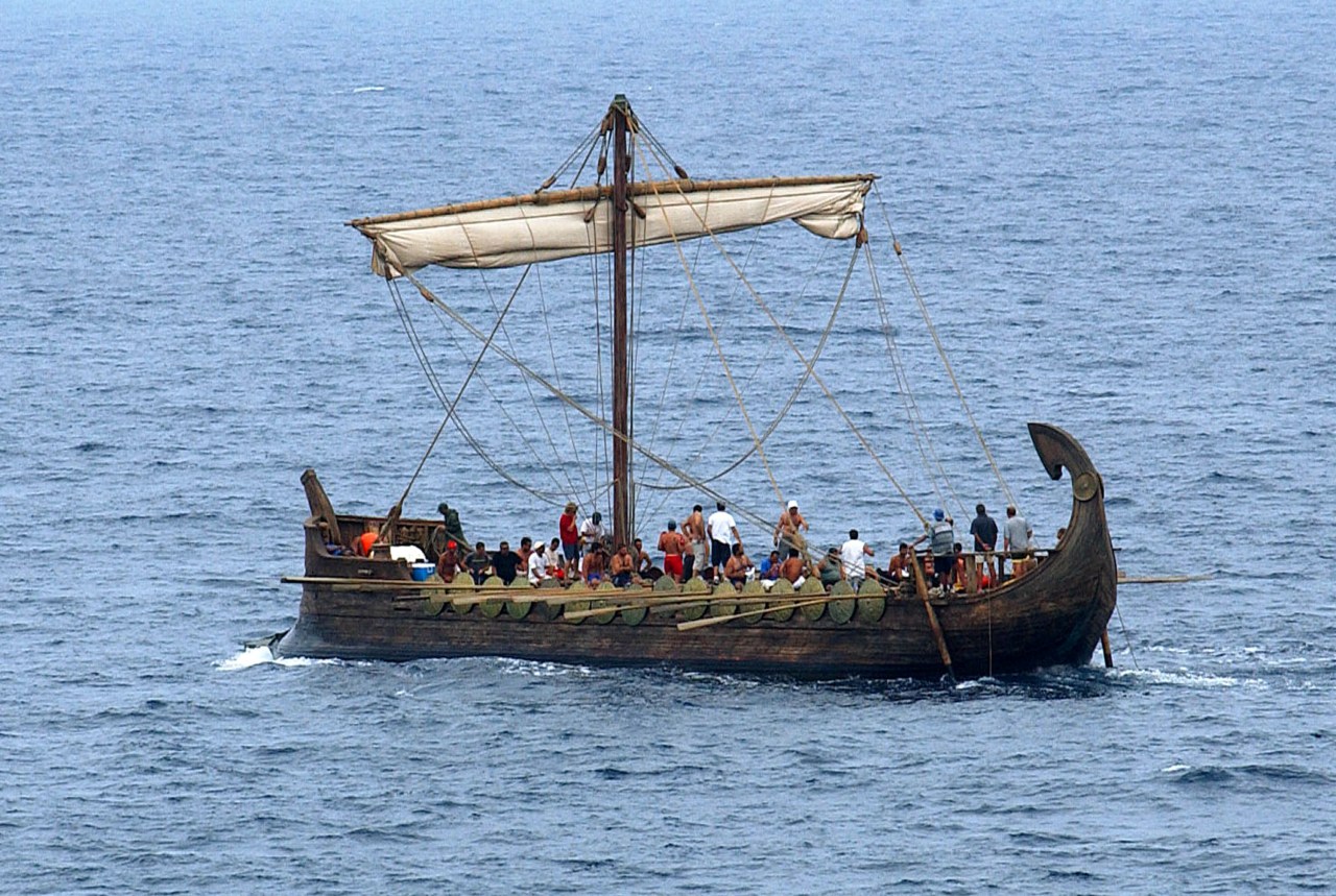 A wooden ancient-style sailing ship with a large mast and white sail on the open sea, filled with crew members on deck.