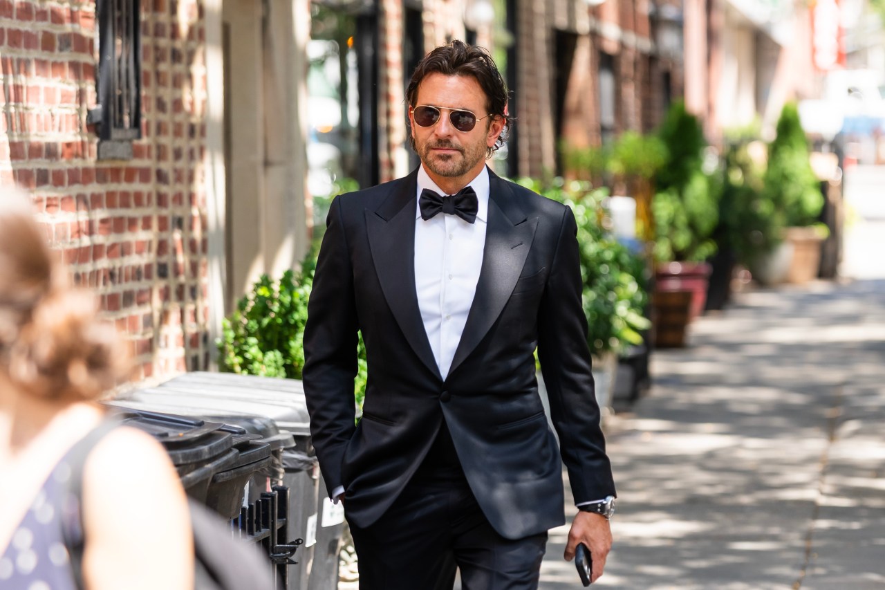 Man in a black tuxedo and bow tie wearing aviator sunglasses walking along a sunlit sidewalk in New York City's West Village.