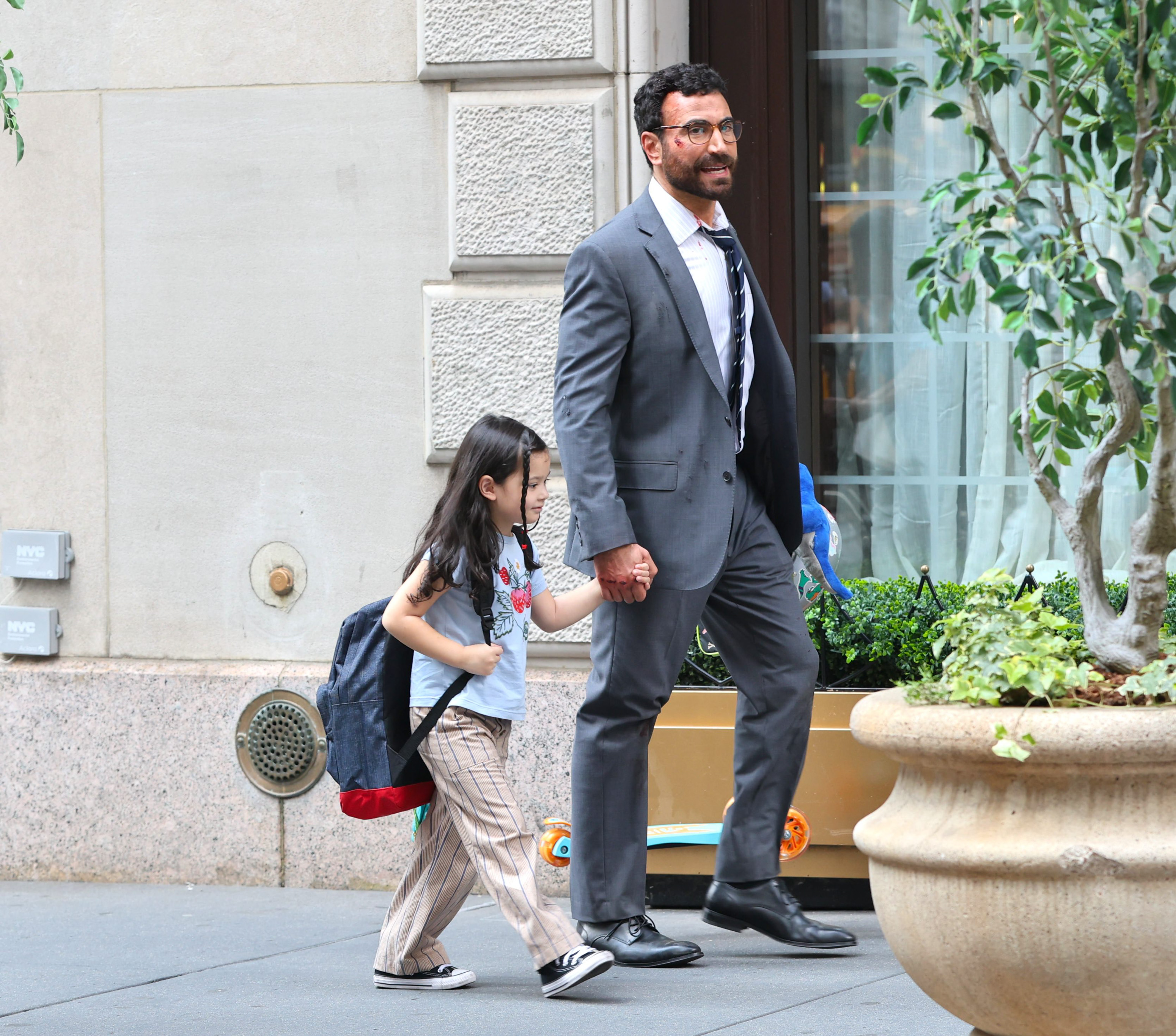Man in gray suit walks briskly holding a young girl's hand outside a stone building; girl wears a backpack and striped pants.