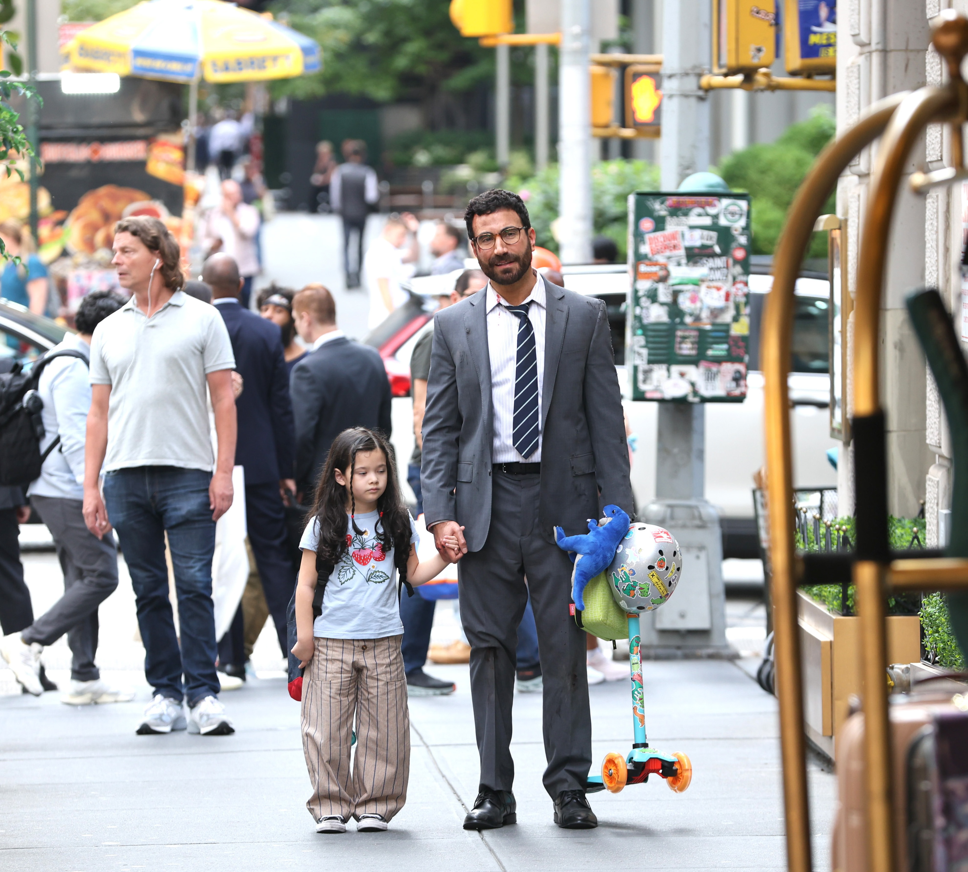 Man in gray suit and tie holds a young girl's hand on a crowded New York City sidewalk while carrying a colorful kids' scooter.