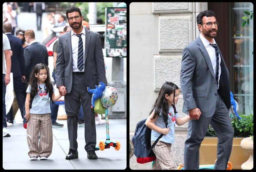 Man in a gray suit and glasses walks hand-in-hand with a young girl carrying a backpack on a city sidewalk.