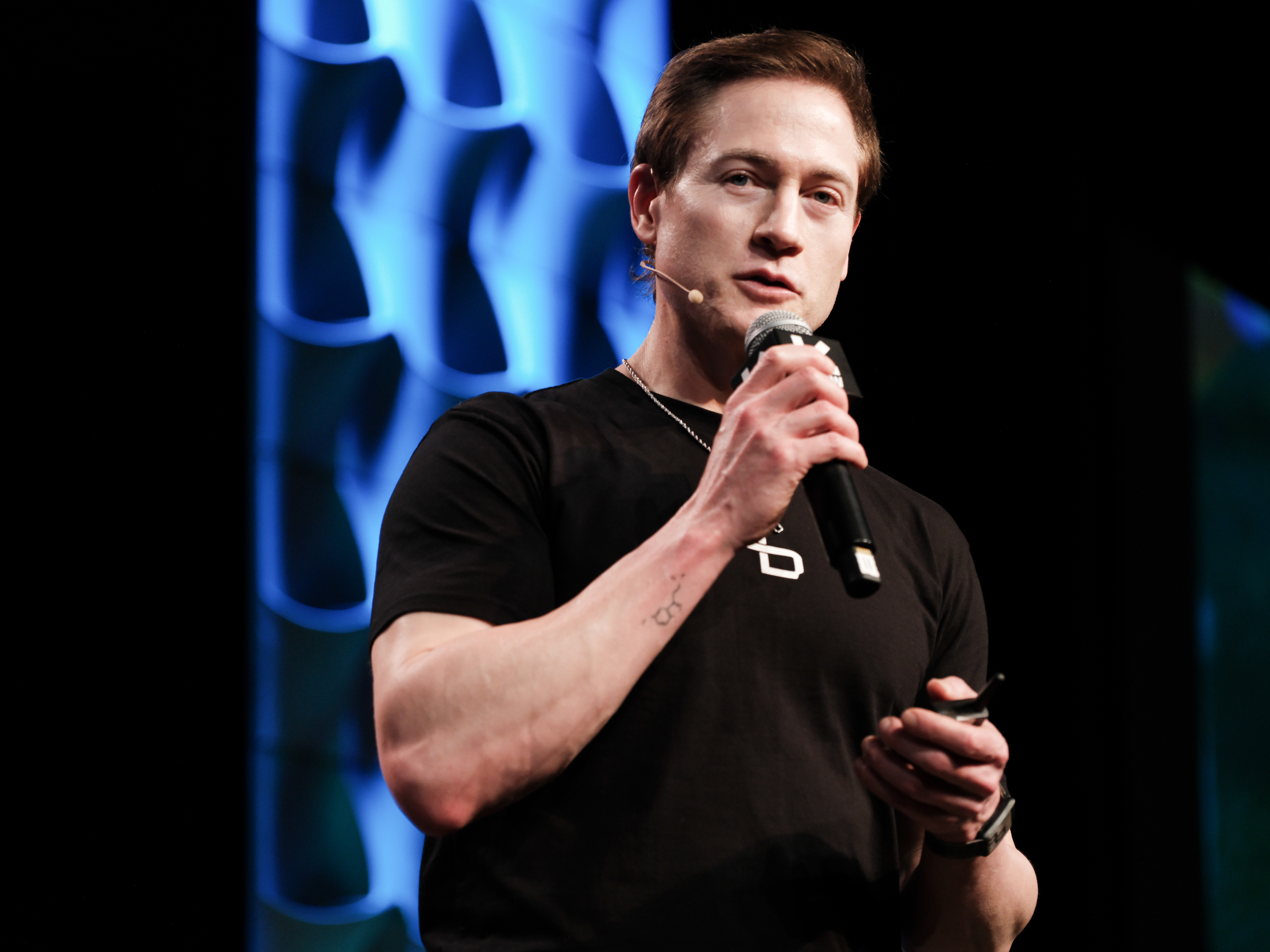 Bryan Johnson in a black t-shirt holding a microphone on stage at SXSW 2025, with a blue illuminated backdrop behind him.