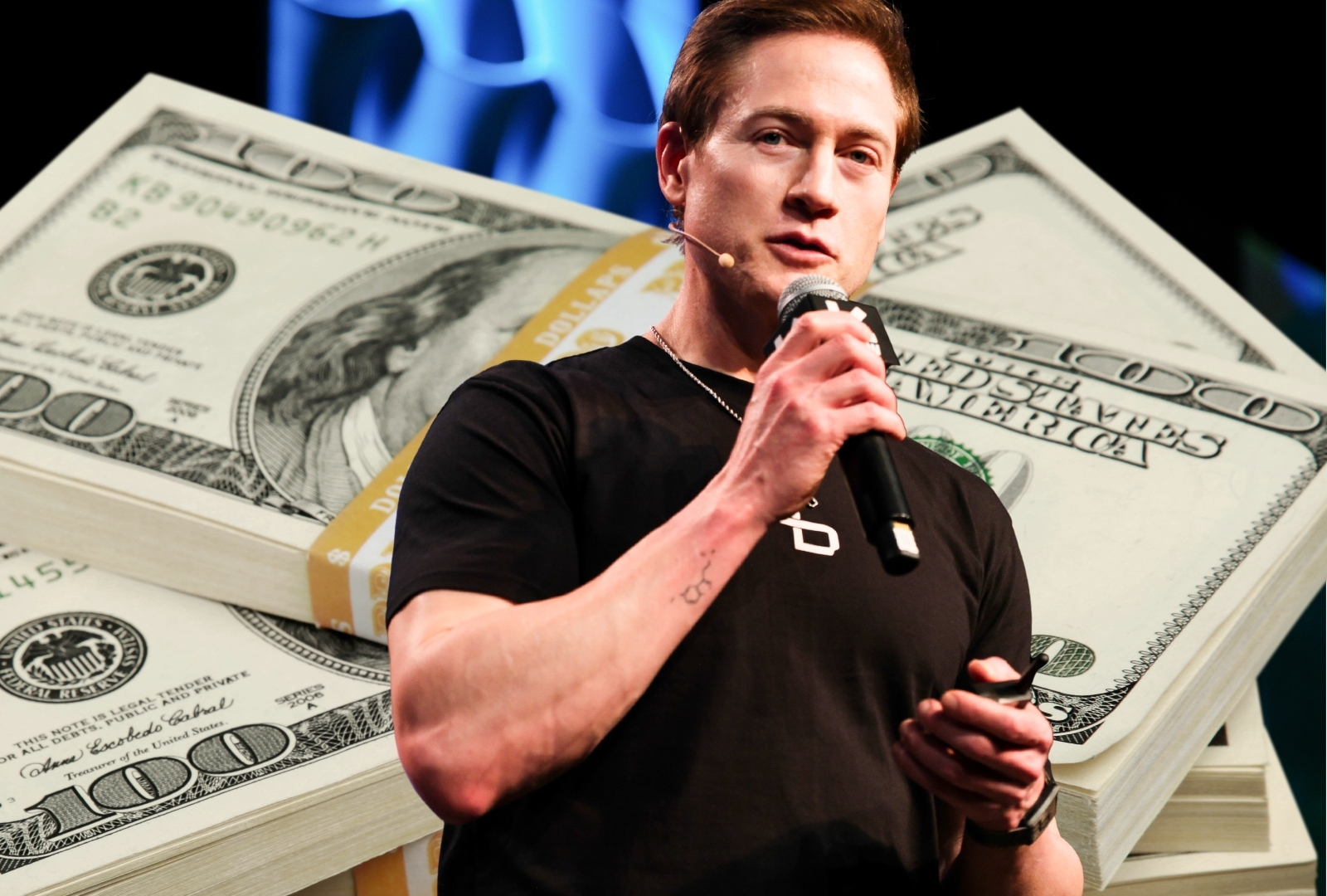 Bryan Johnson speaking on stage into a handheld microphone, wearing a black t-shirt, with a blue lit backdrop behind him.