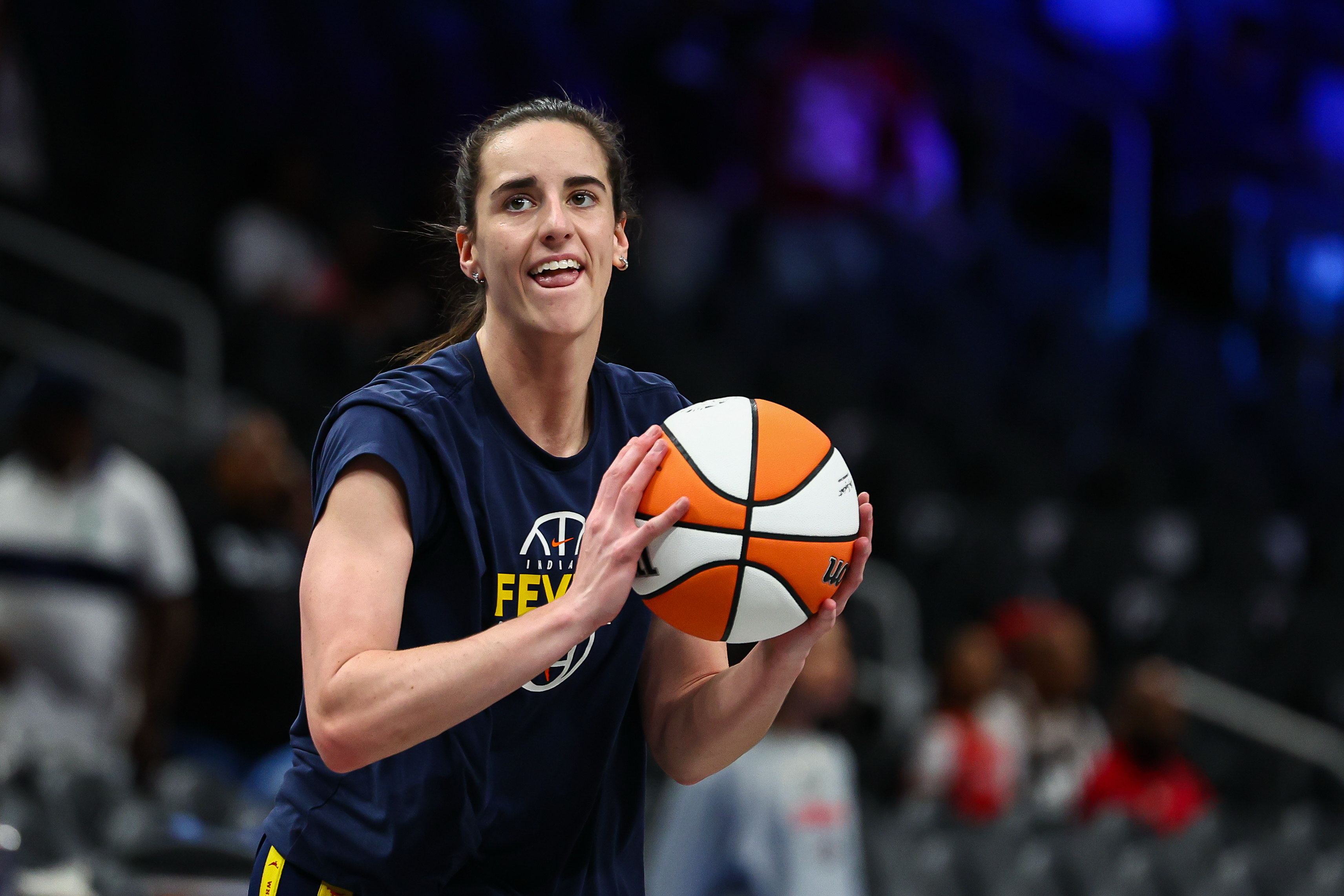 A female basketball player in a navy Indiana Fever jersey holds a basketball in shooting position during pregame warmups in an arena.