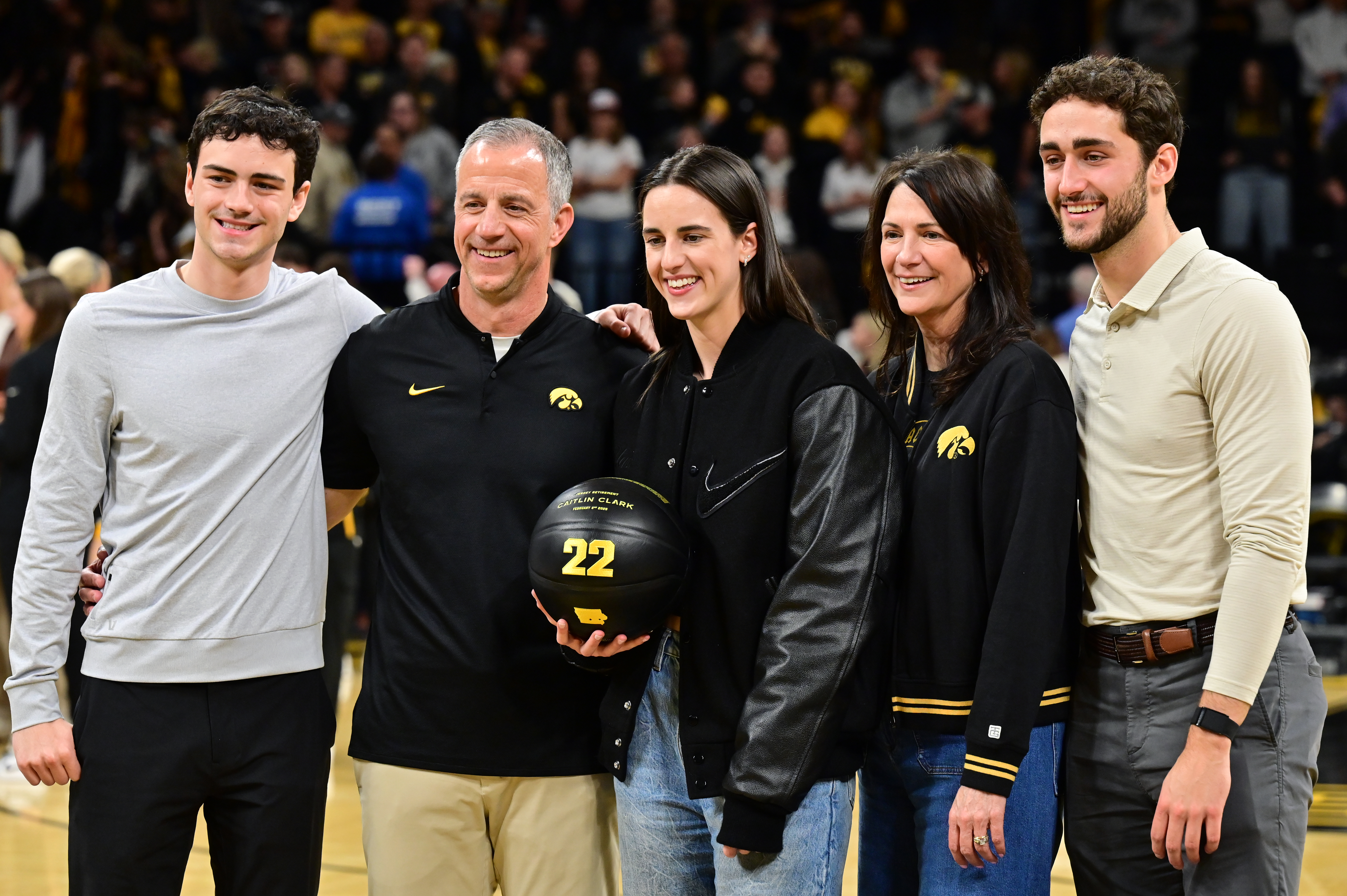 Caitlin Clark poses with four family members on a basketball court, one holding a black commemorative basketball with the number 22 on it.