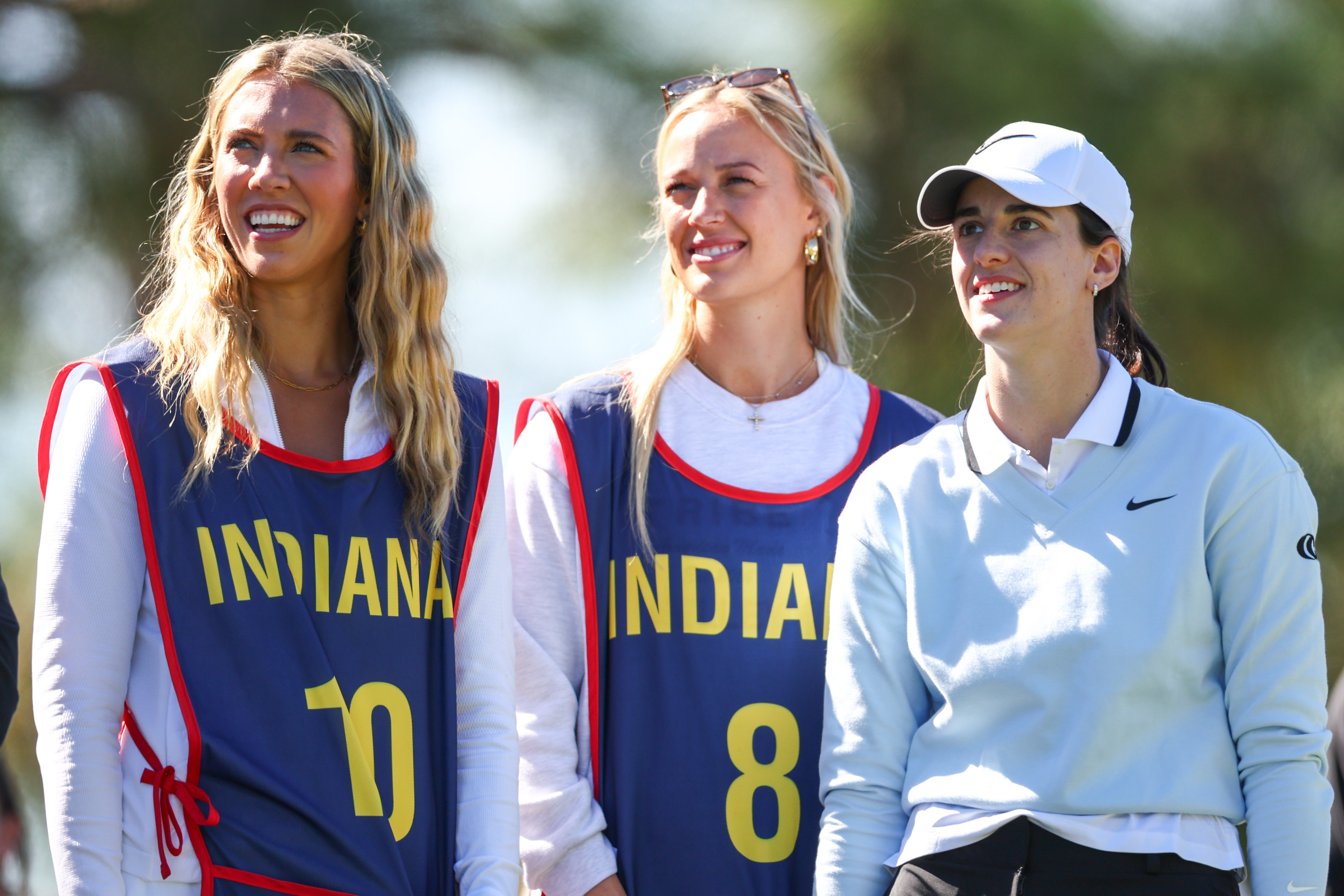 Three women standing outdoors on a golf course; the two on the left wear blue Indiana caddie bibs numbered 10 and 8, while the woman on the right wears a white Nike golf outfit and cap.