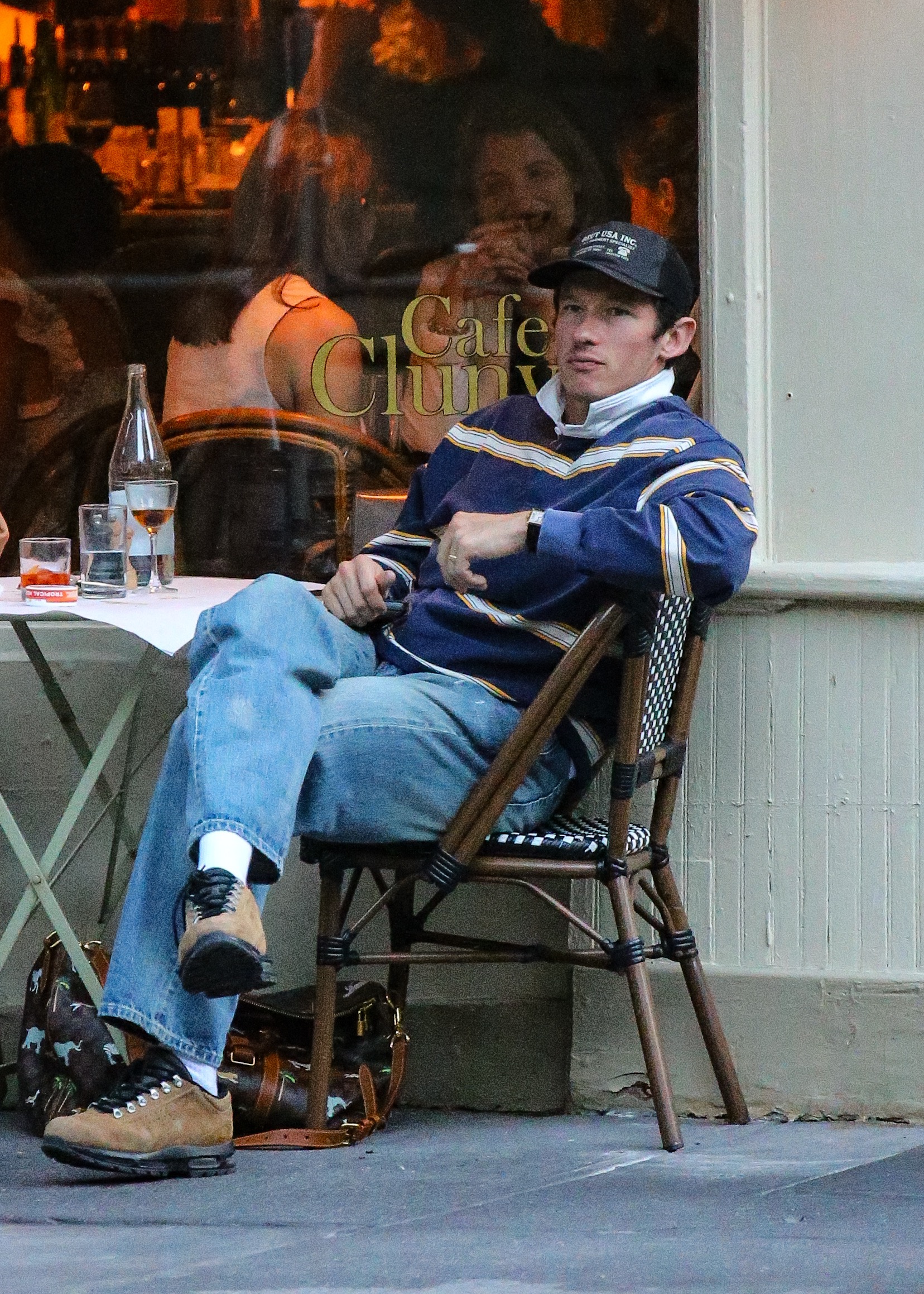 Man in a navy striped sweater and baseball cap sits in a bistro chair outside Cafe Cluny with a patterned duffle bag on the ground.
