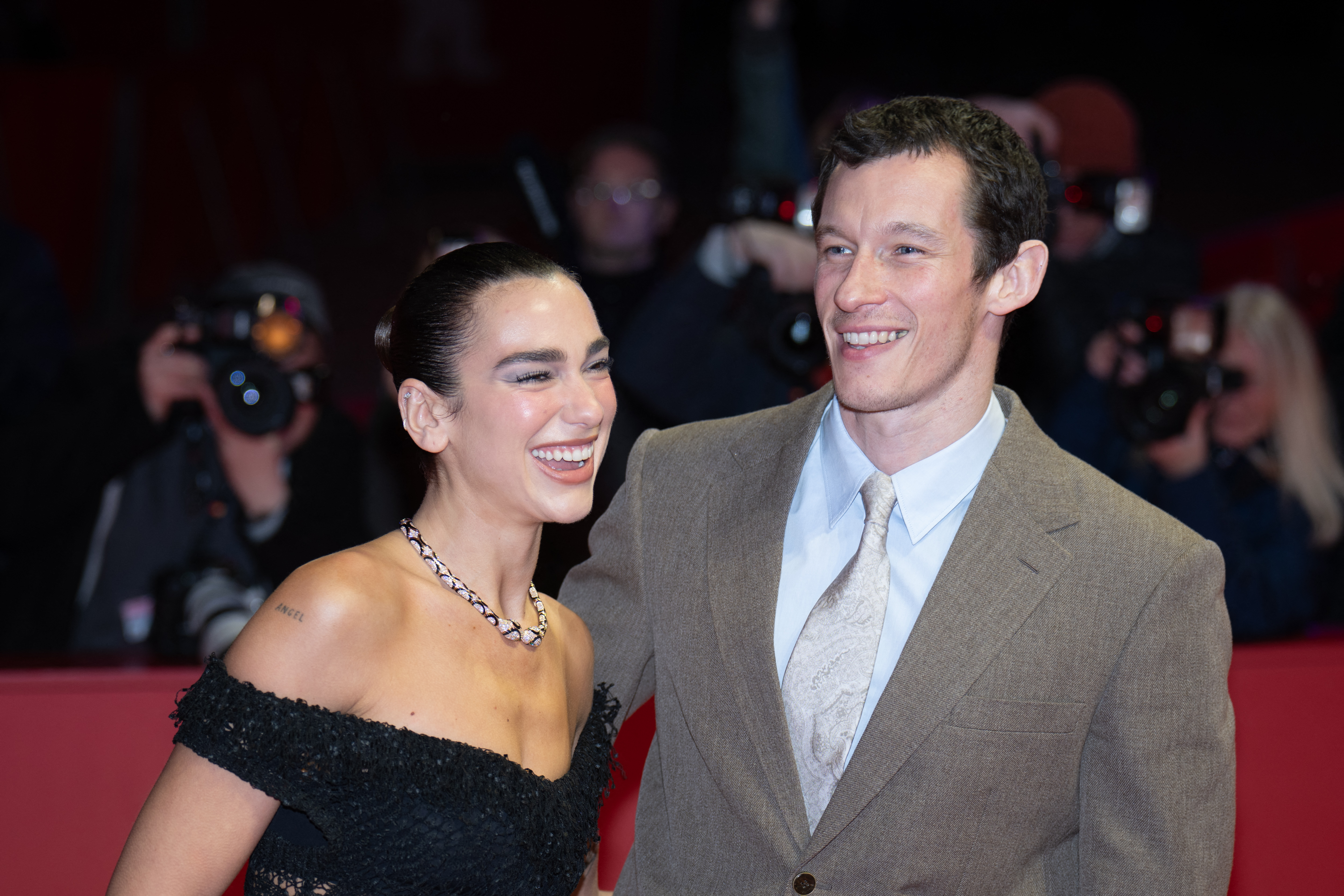 Dua Lipa in a black off-shoulder dress and Callum Turner in a tan suit smiling together on a red carpet with photographers behind them.