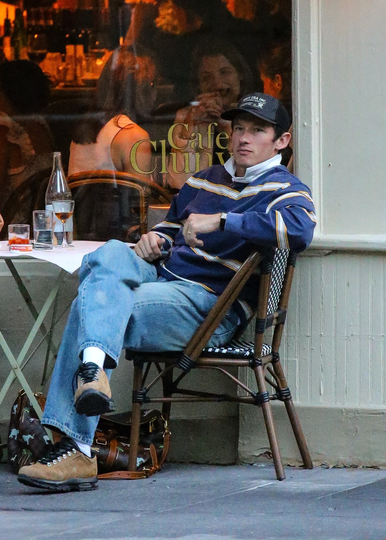 Man in a navy striped sweater and black cap seated in a bistro chair outside Cafe Cluny, with drinks on the table beside him.