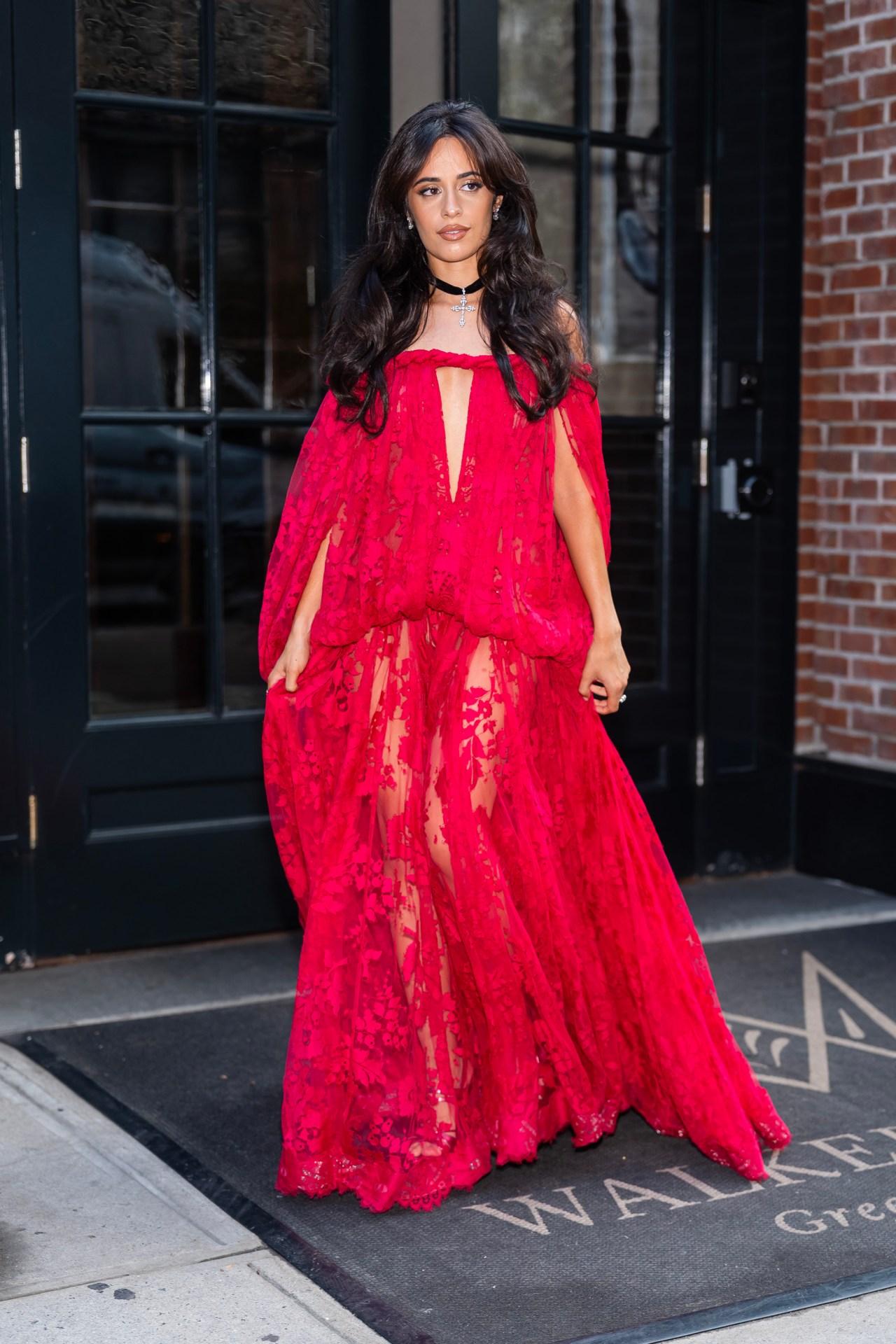 Camila Cabello wearing a sheer red lace floor-length gown with cape sleeves and a black choker necklace, standing outside a building in New York City.