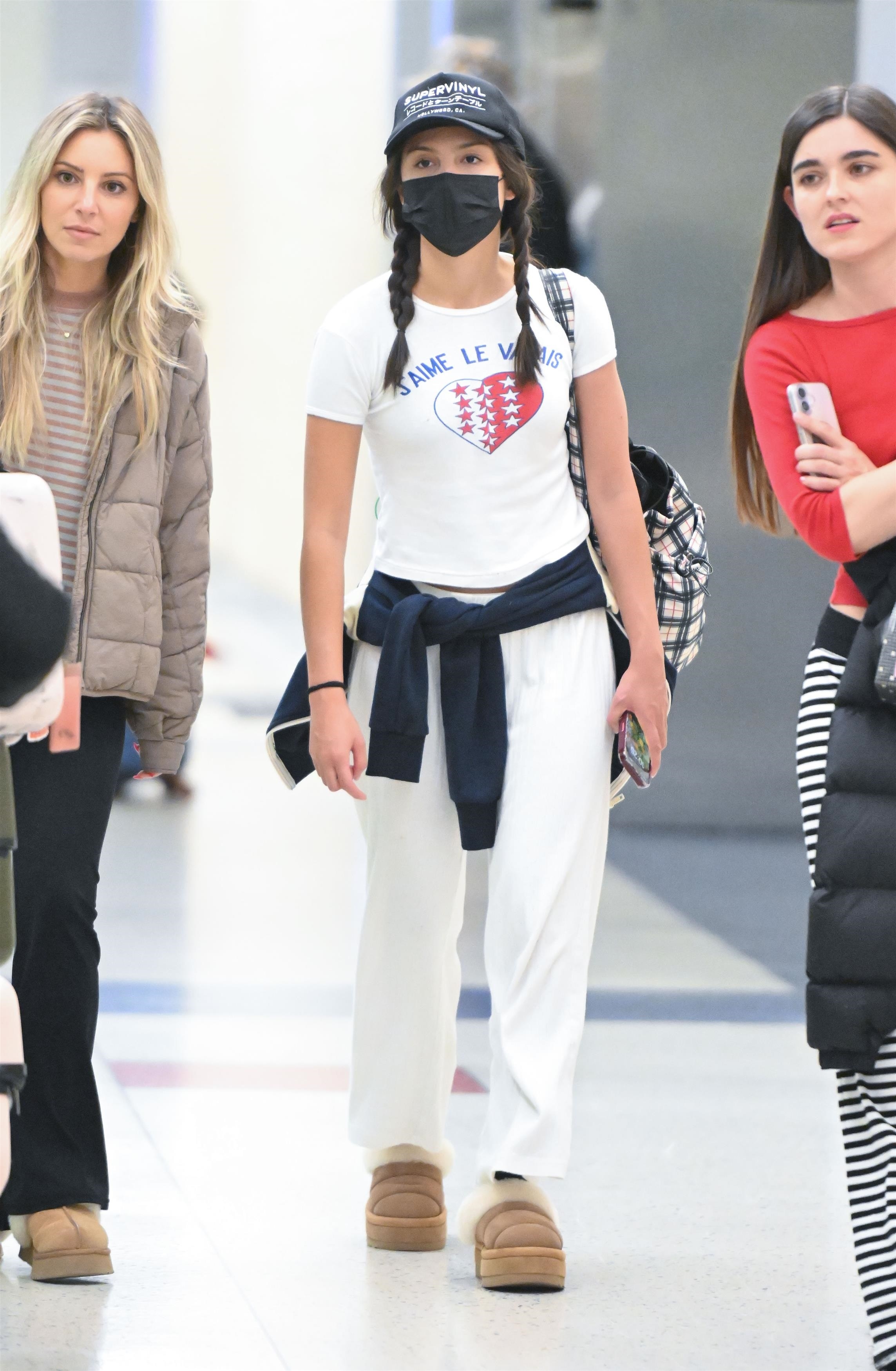 Young woman in black cap, face mask, braids, white graphic tee, white wide-leg pants, and UGG platform sandals walking through an airport terminal.