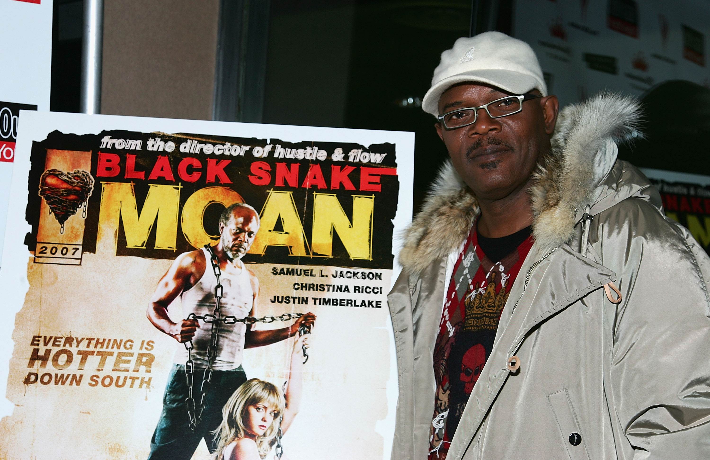 Samuel L. Jackson in a white cap and fur-trimmed coat standing next to a Black Snake Moan movie poster at a premiere.