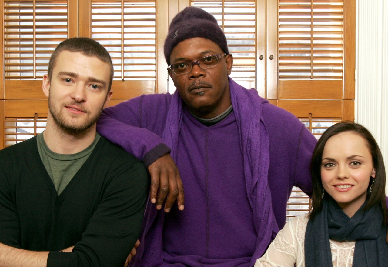 Three people pose together indoors in front of wooden shutters: a man in a black shirt on the left, a taller man in a purple sweater and beanie in the center, and a woman in a white top and blue scarf on the right.