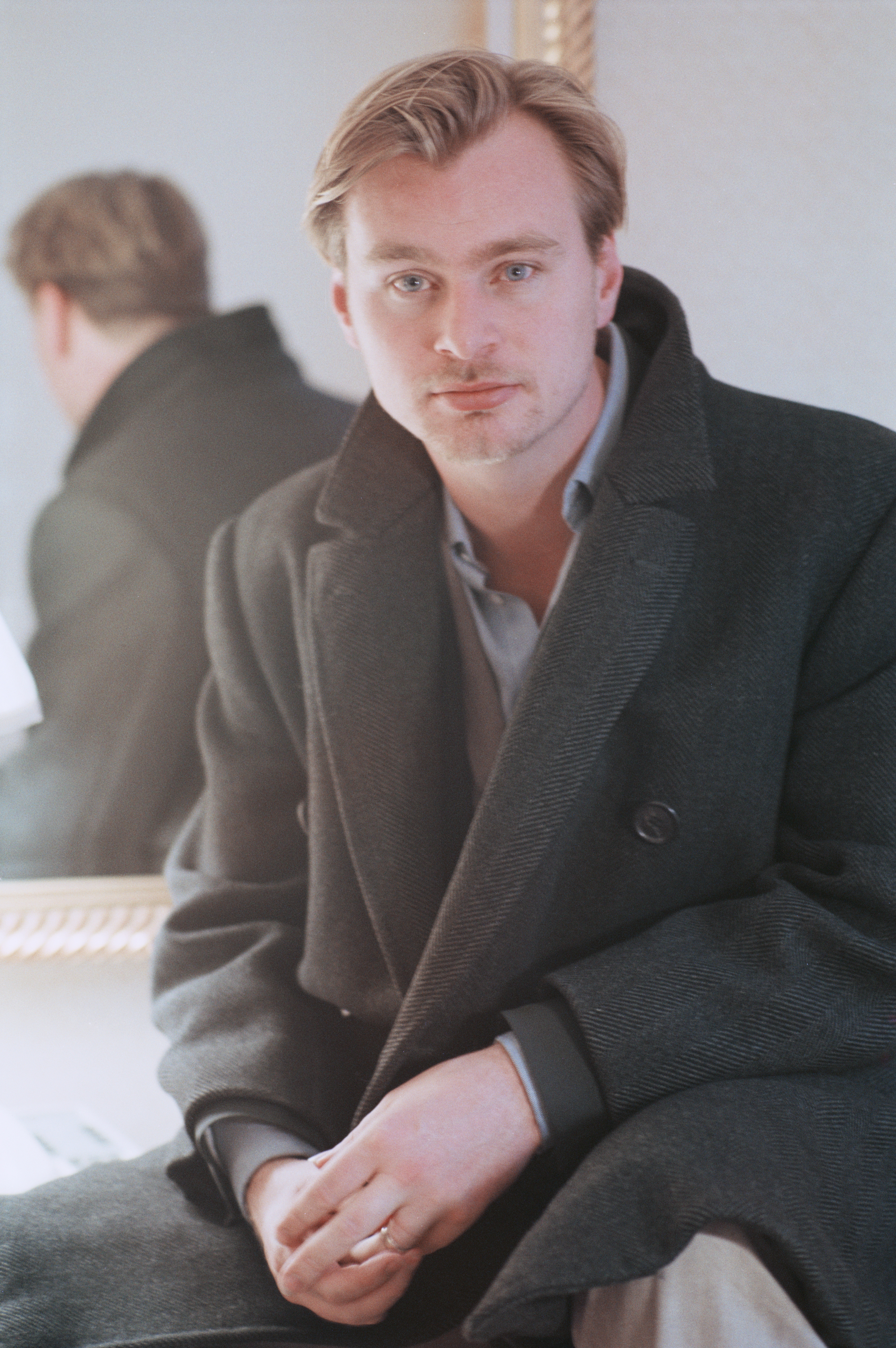 Young Christopher Nolan seated, wearing a dark overcoat over a grey shirt, hands clasped, looking at the camera.