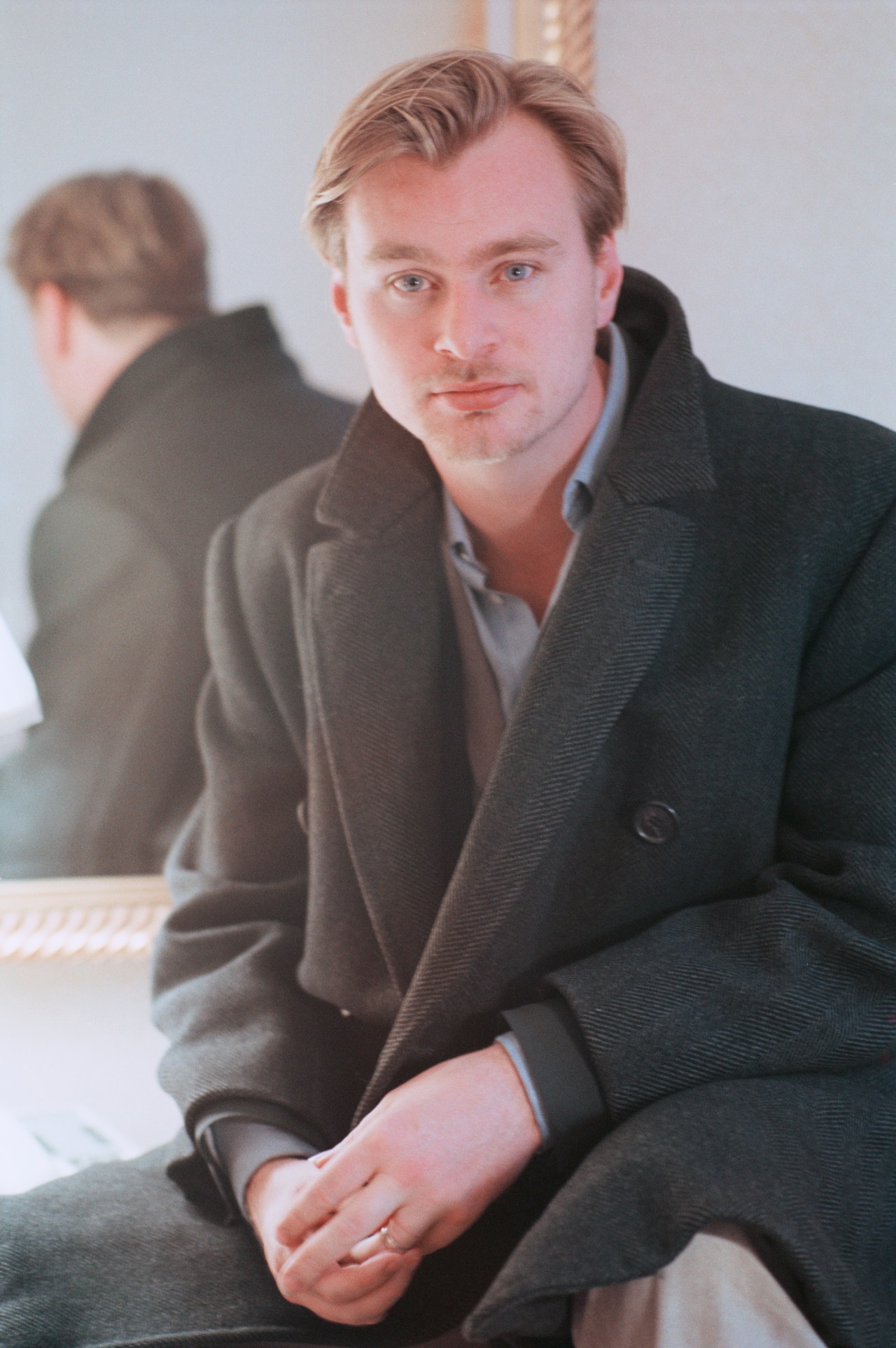 Young Christopher Nolan seated, wearing a dark overcoat over a grey shirt, hands clasped, looking at the camera.