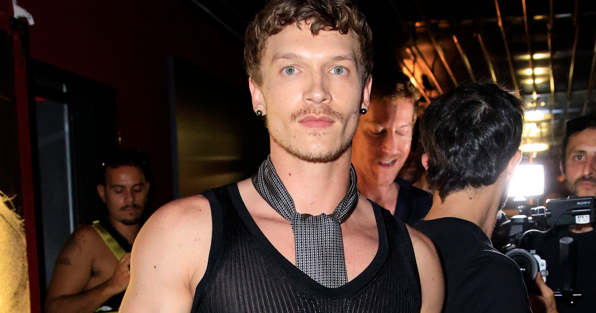 Connor Storrie in a sheer black ribbed tank top and chain necklace at an event in Paris, with photographers visible in the background.