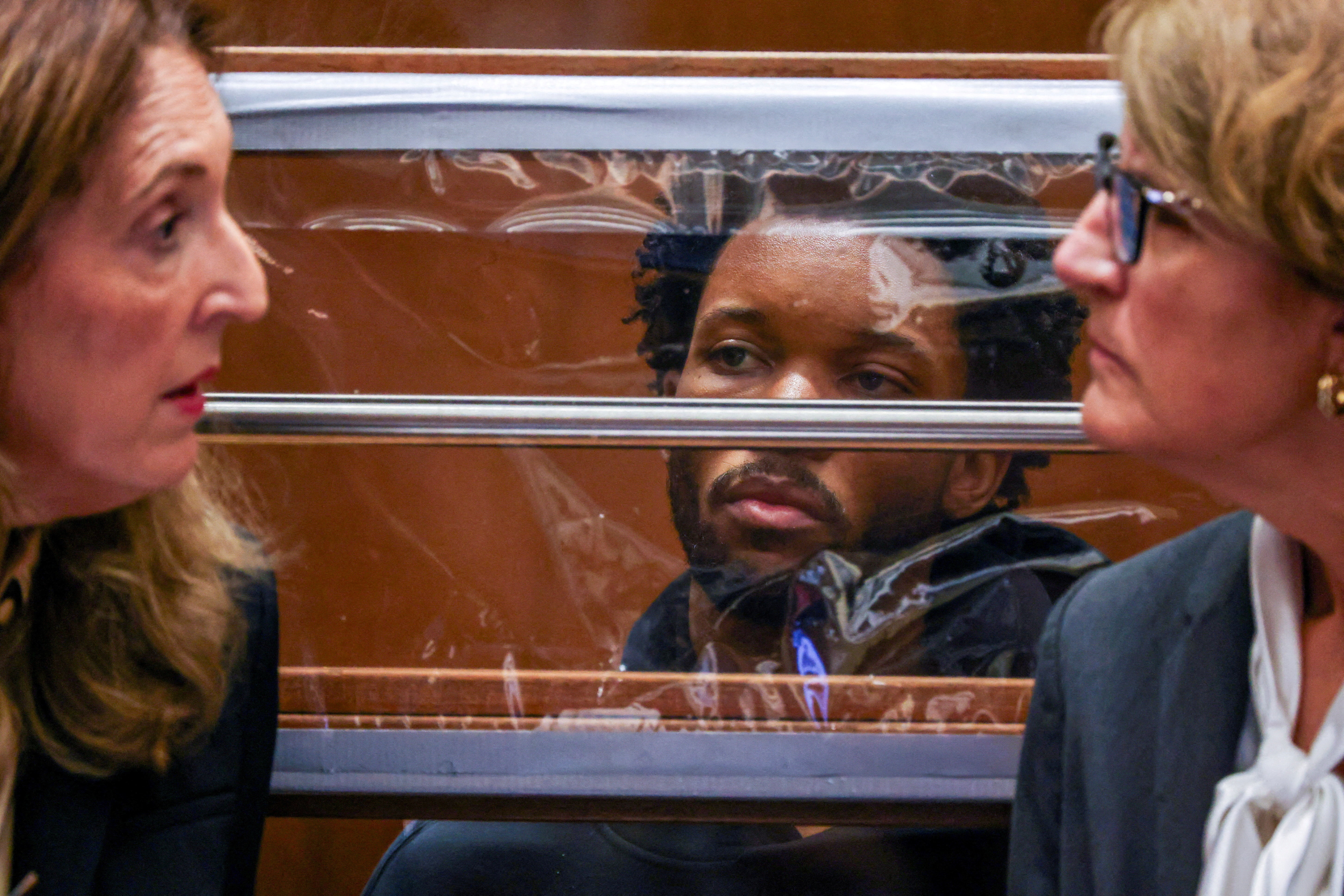 A man seen behind a glass partition between two women attorneys in a courtroom; one attorney speaks while the other looks on.