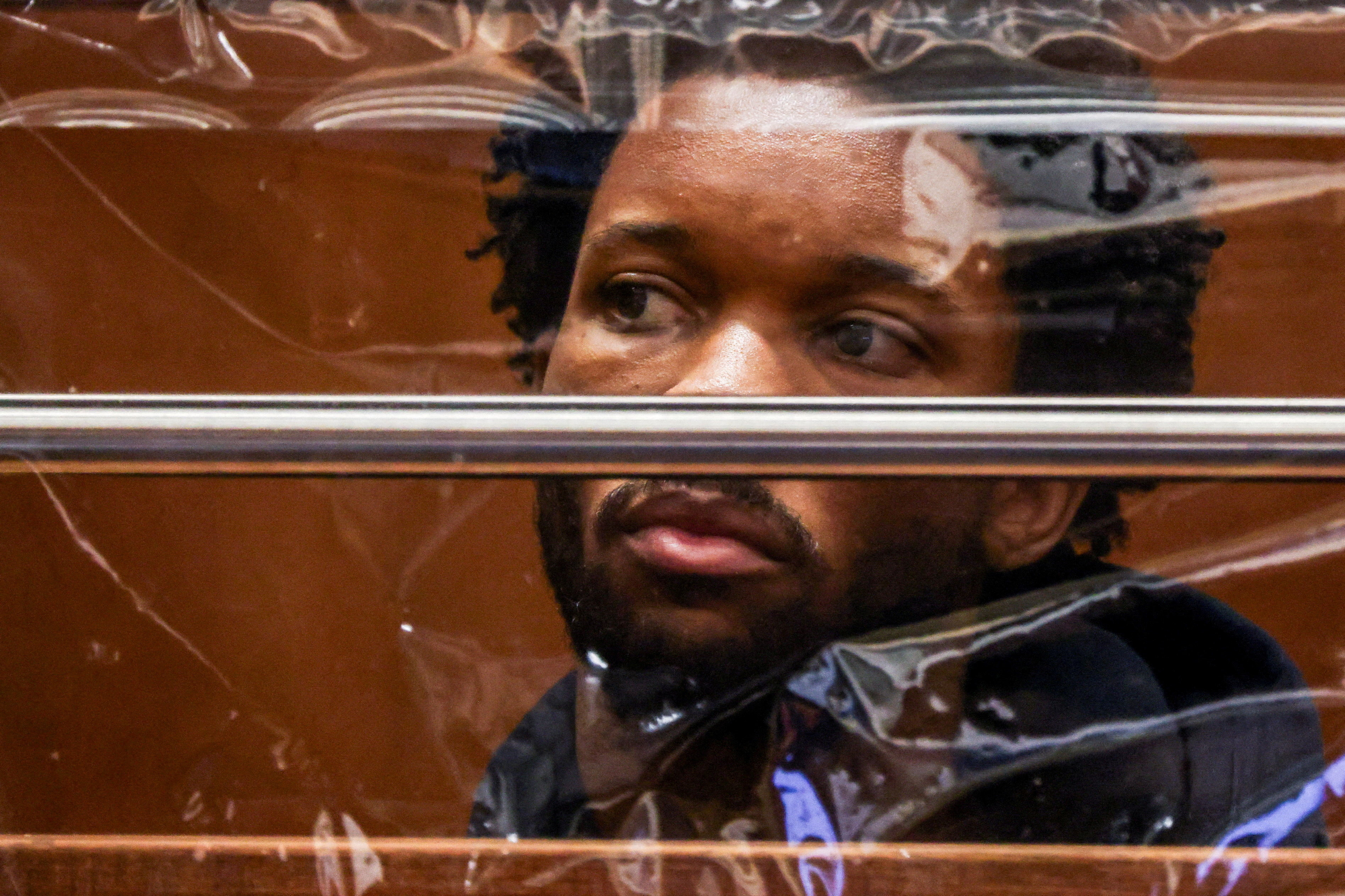 A man with short braids and a beard looks sideways through a scratched glass partition with metal bars in a courtroom setting.