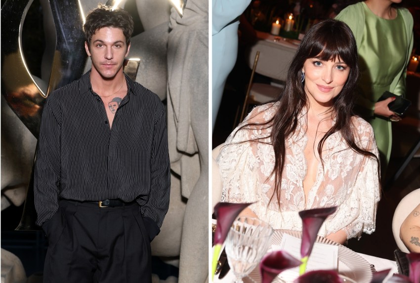 Man in a black pinstripe shirt and wide-leg black trousers standing in front of stone sculptures and a gold YSL logo at a fashion event.