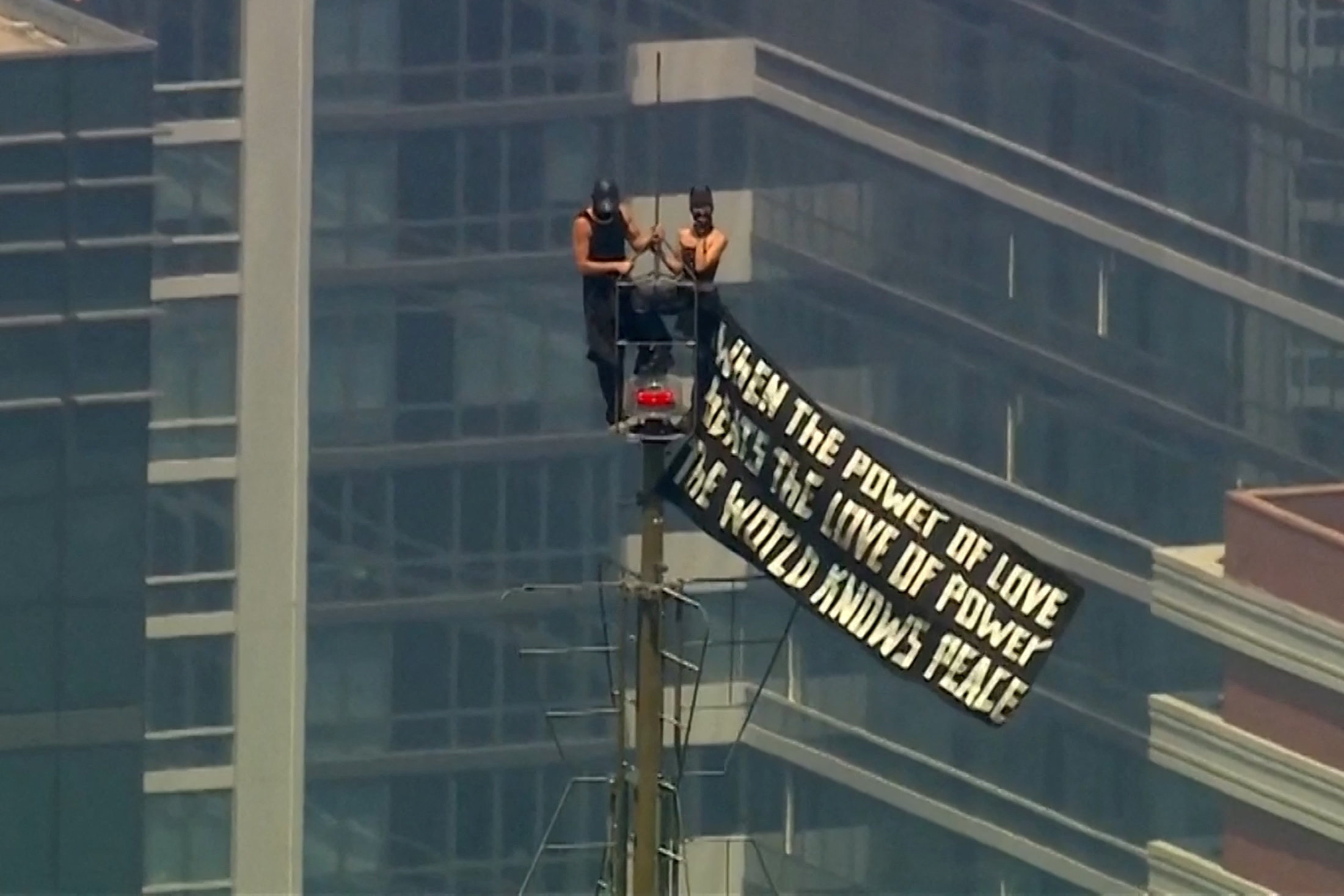 Two masked people stand on a ledge near the top of the Empire State Building holding a large banner with a peace quote, with Manhattan skyscrapers behind them.