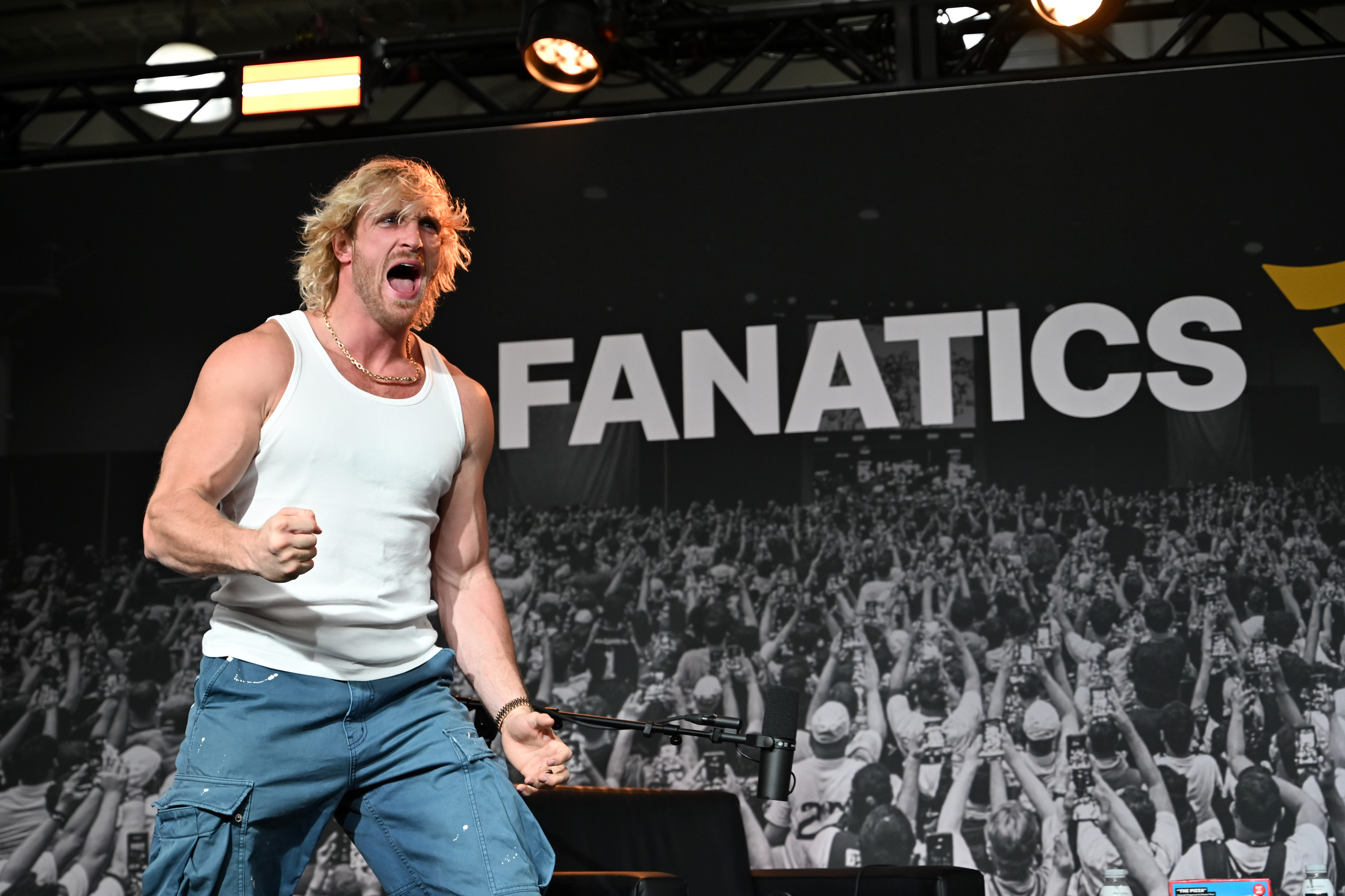Logan Paul in a white tank top and blue cargo pants shouting and pumping his fist onstage in front of a Fanatics banner.