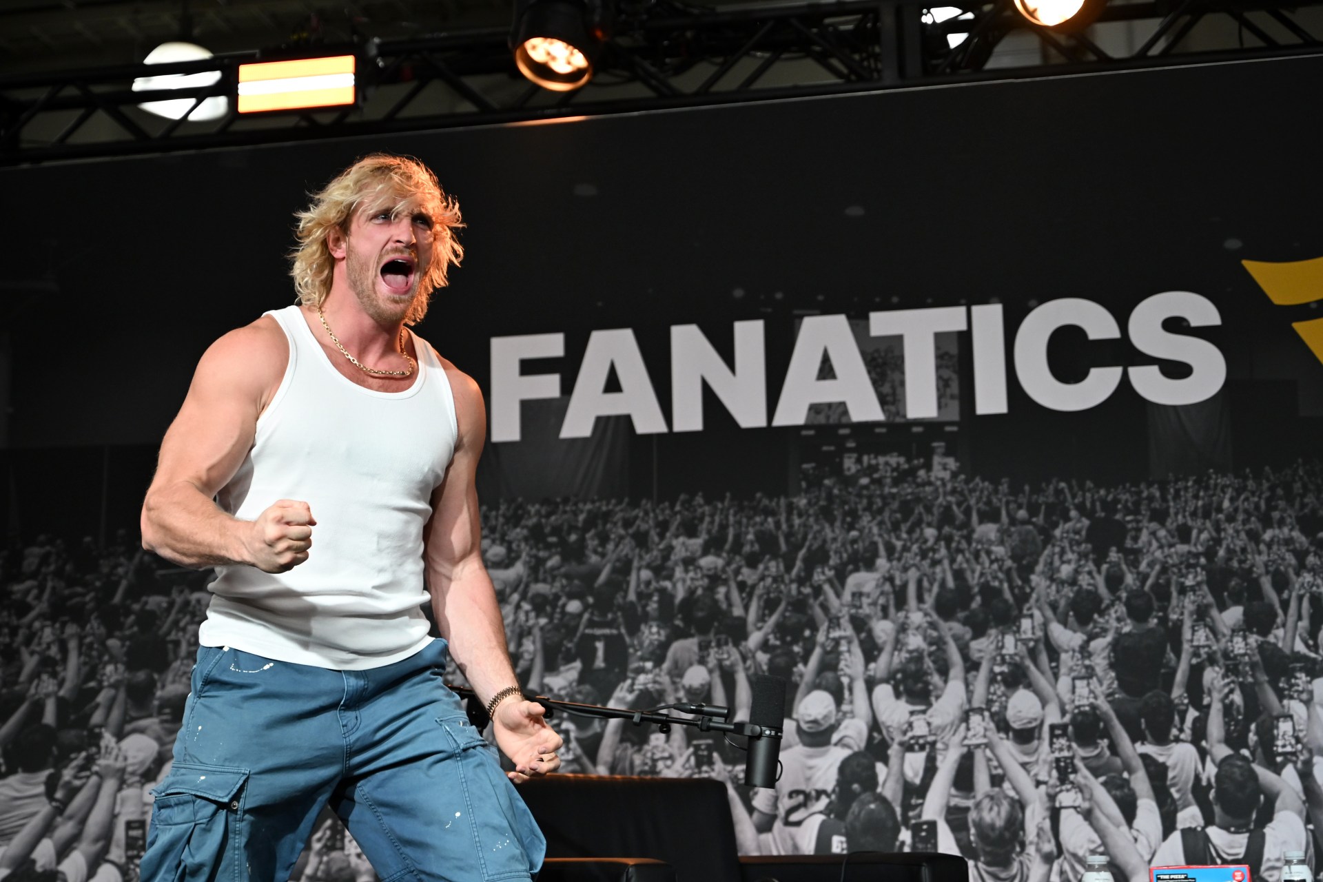Logan Paul in a white tank top and blue cargo pants shouting and pumping his fist onstage in front of a Fanatics banner.