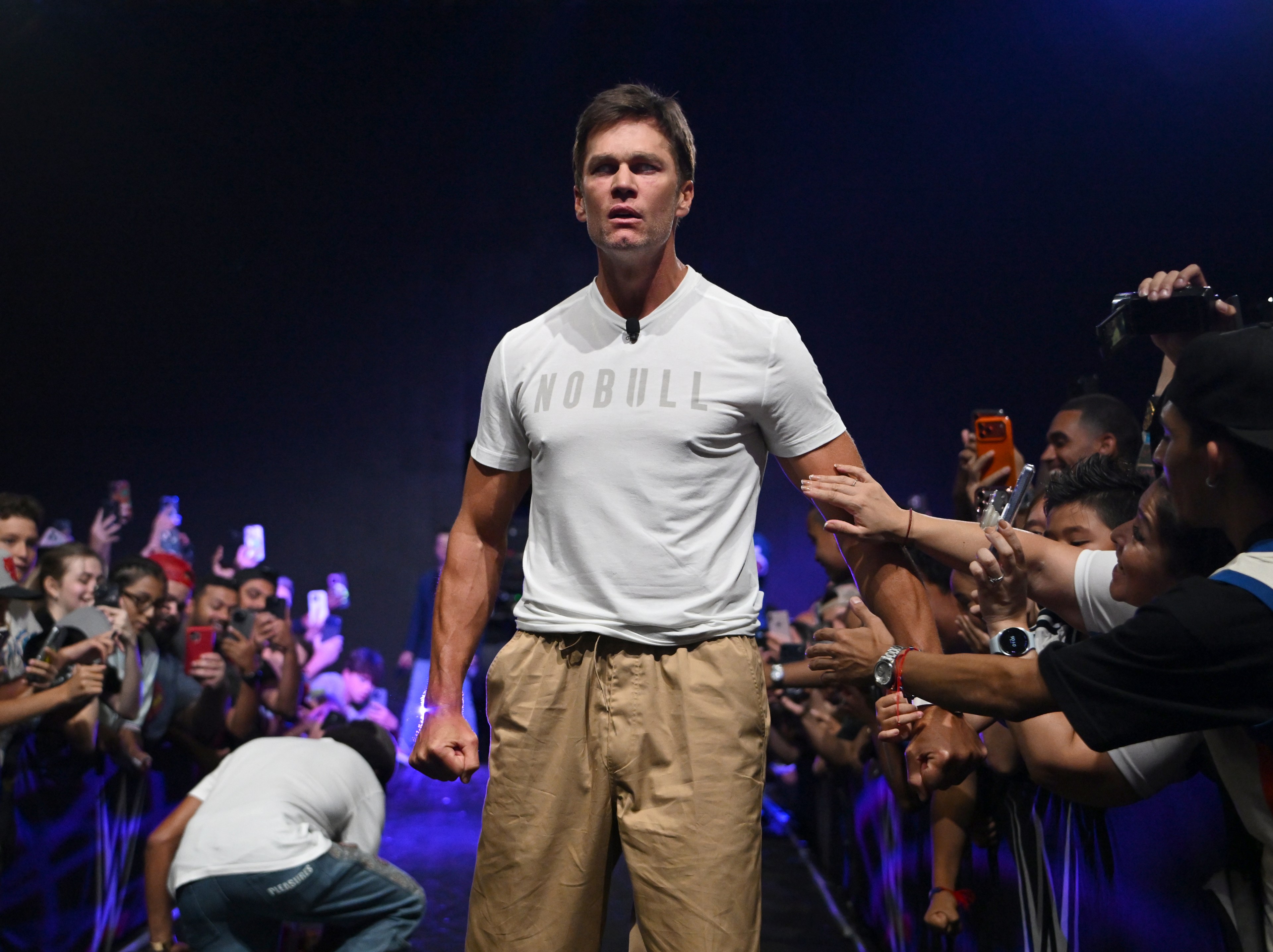 Tom Brady in a white NOBULL t-shirt and khaki pants, fist clenched, walking through an enthusiastic crowd at a large indoor event.