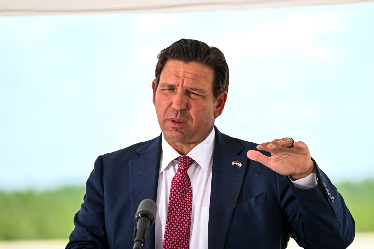 Florida Governor Ron DeSantis in a navy suit and red tie speaking at a podium outdoors, gesturing with one hand.