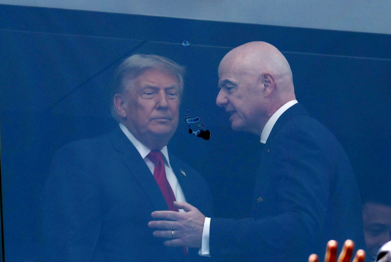 Two men in dark suits talking closely inside a glass-enclosed stadium VIP box, with the football pitch reflected in the window.