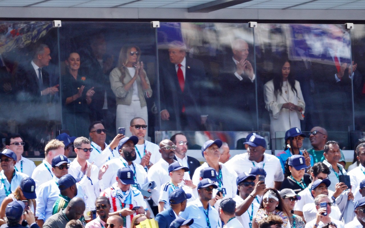 Several people applauding inside a glass-enclosed stadium suite, with a large crowd of attendees in caps and lanyards visible below.