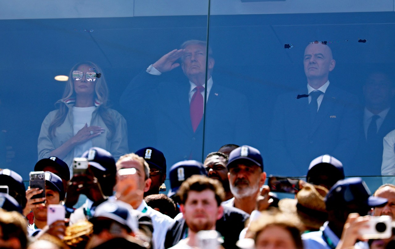 Woman with hand over heart, man in red tie saluting, and bald man in FIFA suit standing behind glass in a stadium VIP box, crowd below photographing them.