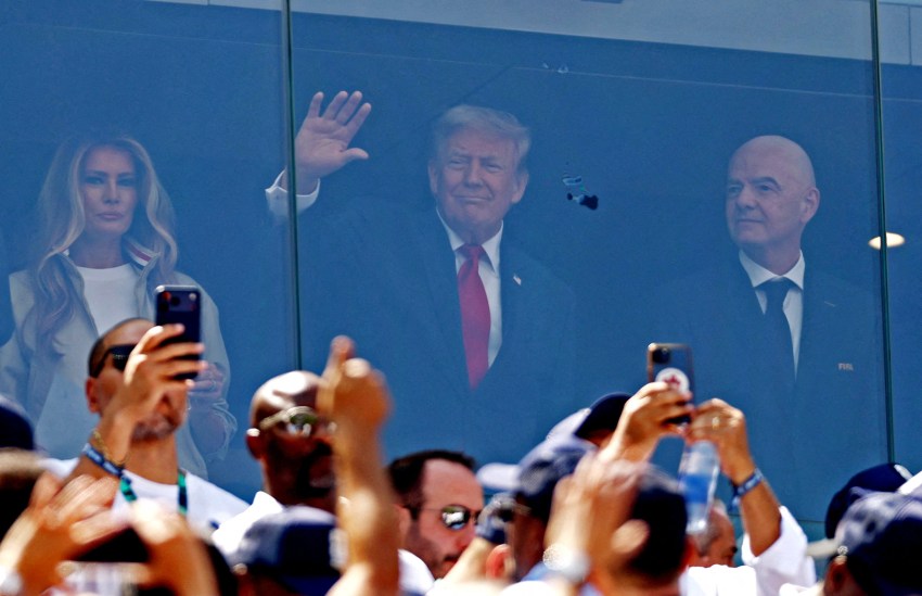 A man in a suit and red tie waves from a glass-enclosed stadium suite while a blonde woman and a bald man stand beside him, as fans below photograph them.