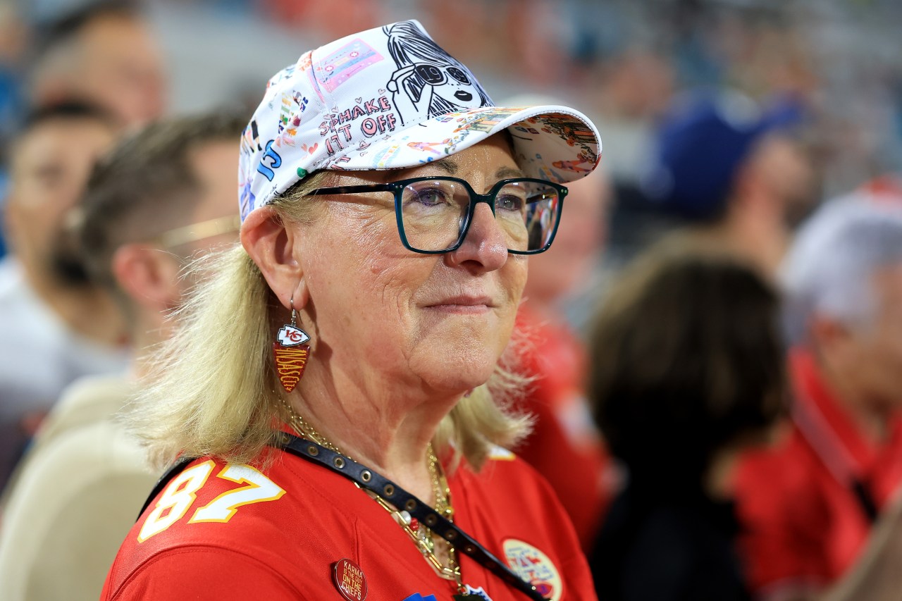 Donna Kelce wearing a red Chiefs #87 jersey, Kansas City Chiefs earrings, and a colorful Taylor Swift-themed cap at a stadium.