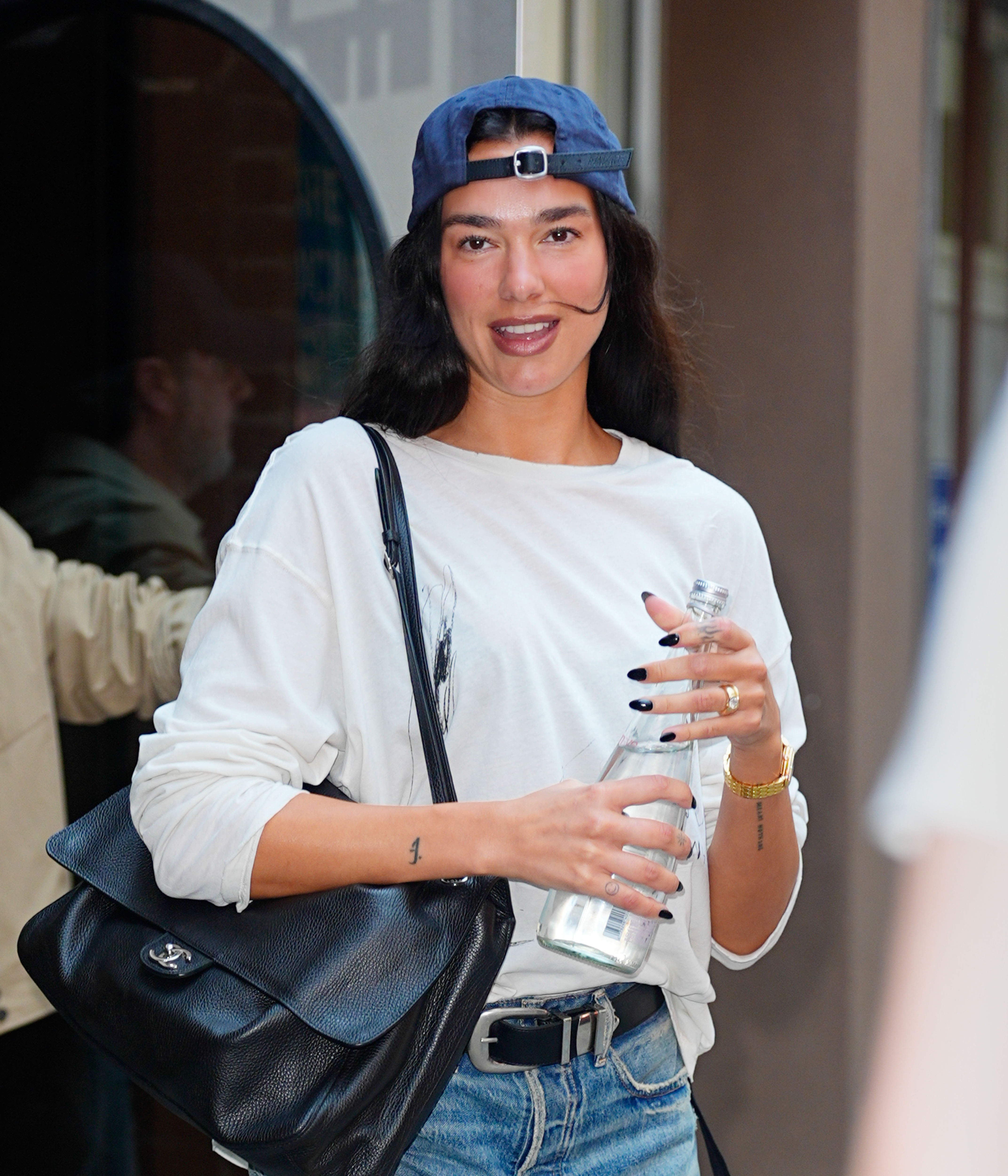 Dua Lipa wearing a backwards blue baseball cap, white long-sleeve shirt, and denim shorts, holding a glass water bottle and a black leather bag.