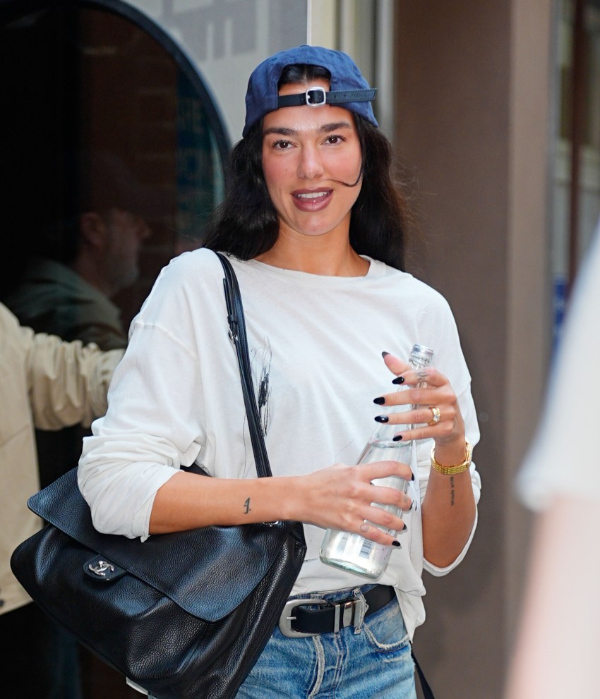 Dua Lipa smiling outdoors in NYC, wearing a backwards blue baseball cap, white long-sleeve shirt, jeans, and a black leather shoulder bag, holding a glass water bottle.