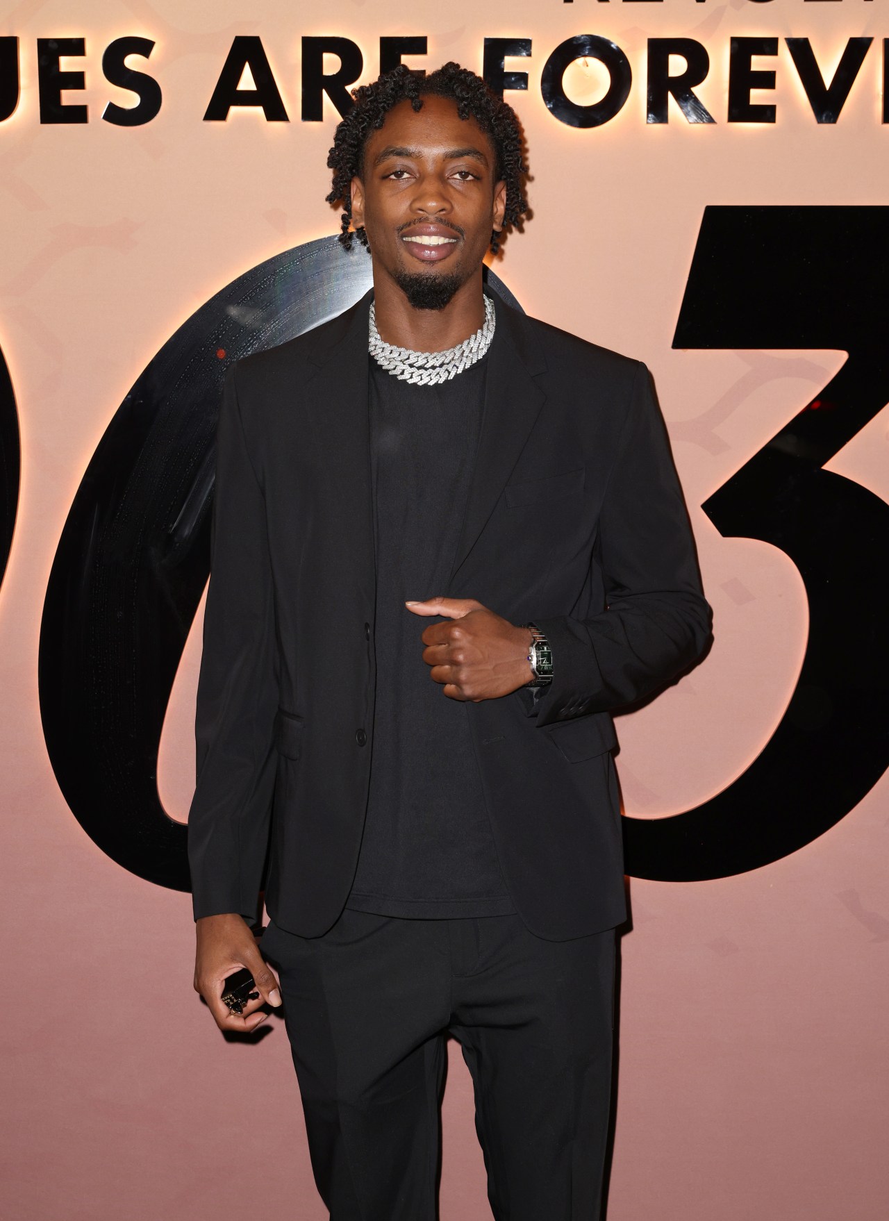 Zaire Wade in a black suit with silver chain necklace and watch, posing at an event with a pink backdrop reading 'Statues Are Forever.'