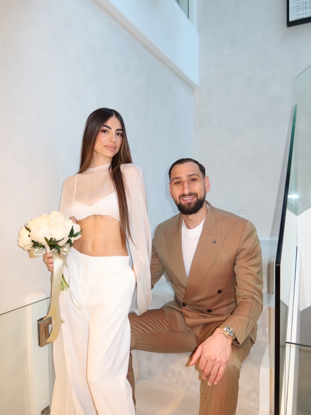 Woman in a sheer white long-sleeve bridal crop top and wide-leg trousers holds a white peony bouquet next to a bearded man in a tan suit on a staircase.