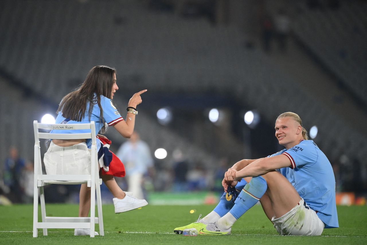 Erling Haaland sitting on a stadium pitch in his Man City kit smiling up at Isabel Haugseng Johansen, who sits on a chair pointing at him.