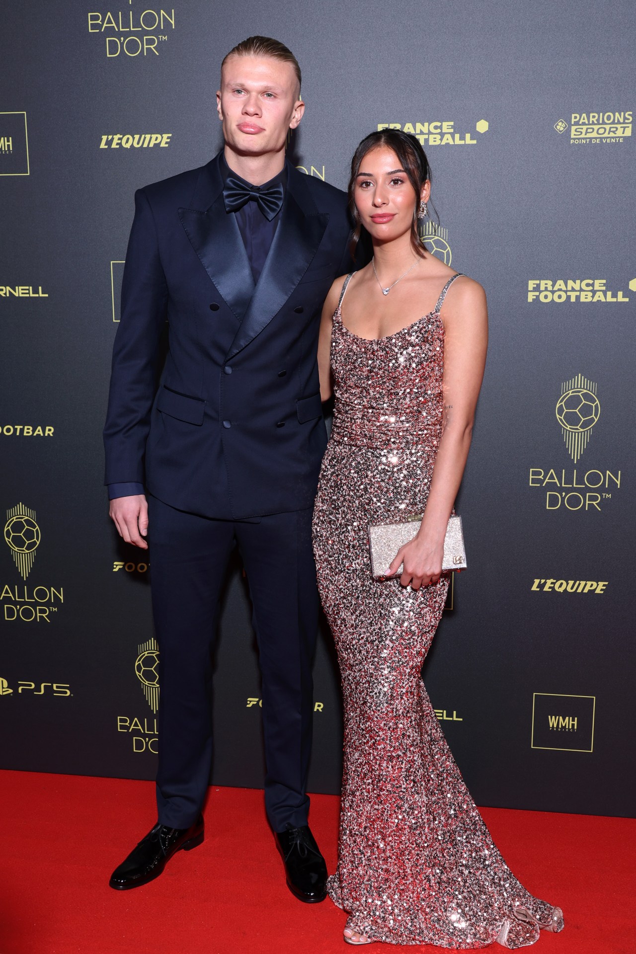 Erling Haaland in a navy tuxedo and Isabel Haugseng Johansen in a sequined gown posing at the Ballon d'Or photocall backdrop in Paris.