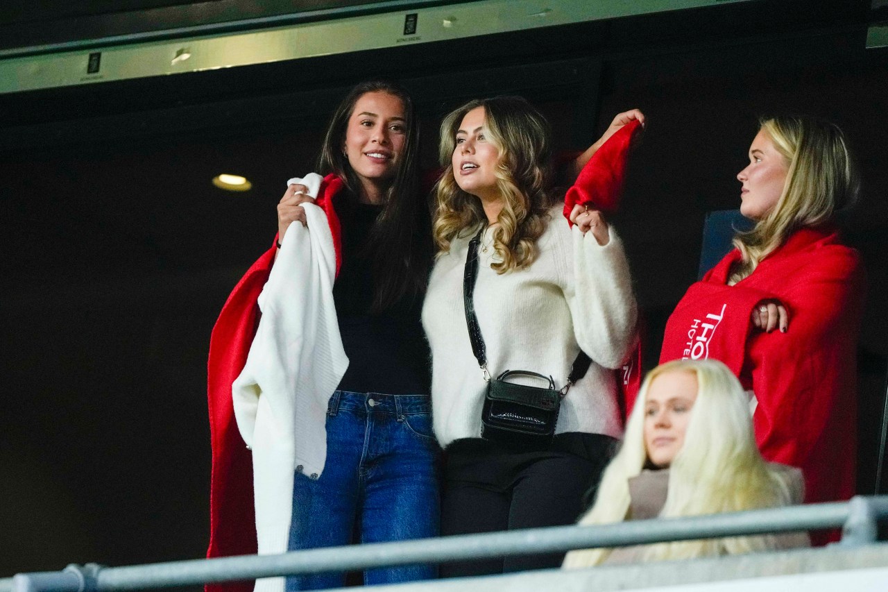 Three young women standing in a stadium VIP box, one holding a white and red blanket, watching a football match.