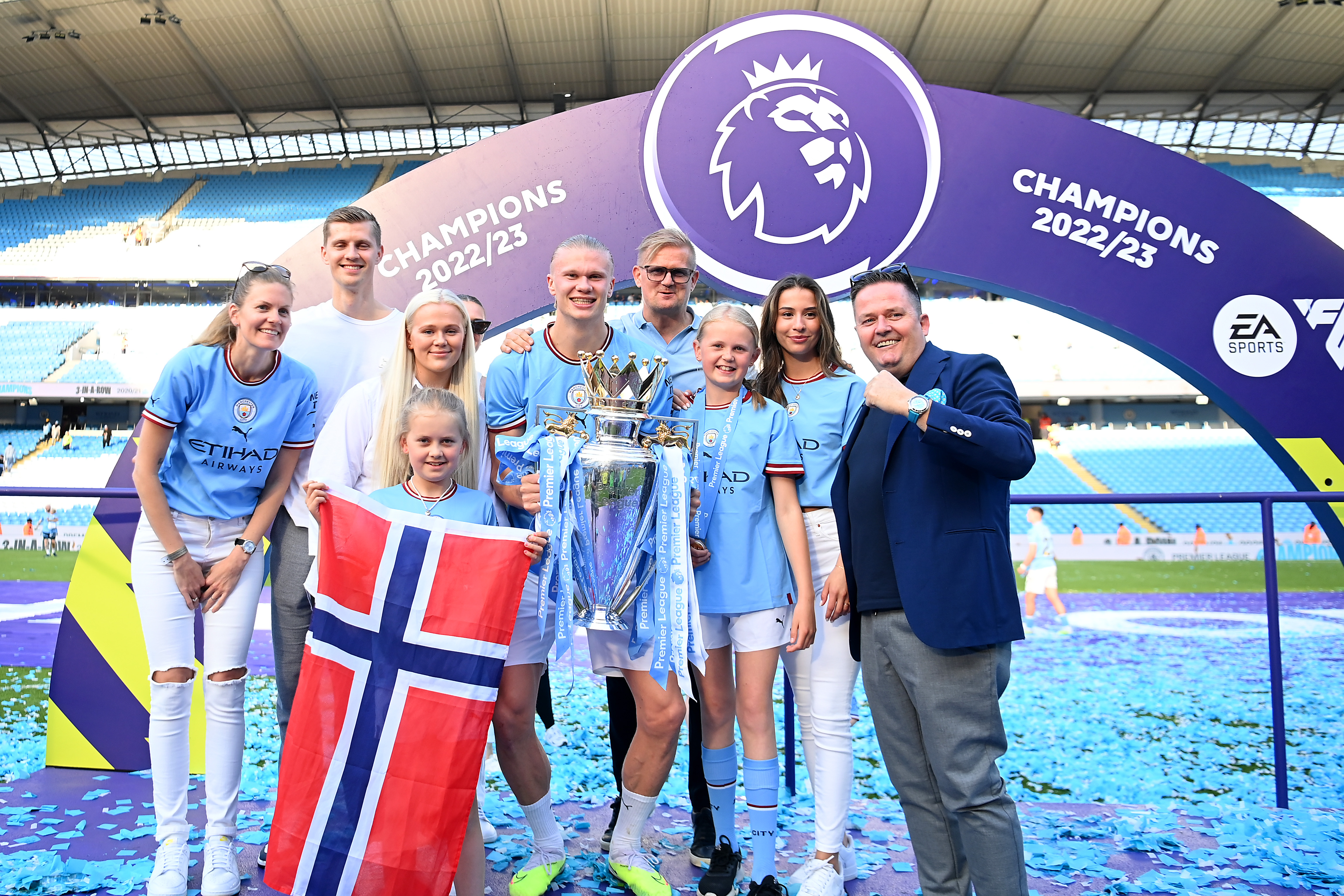 Erling Haaland in a Man City kit holds the Premier League Trophy surrounded by family, one holding a Norwegian flag, under a Champions 2022/23 arch at Etihad Stadium.